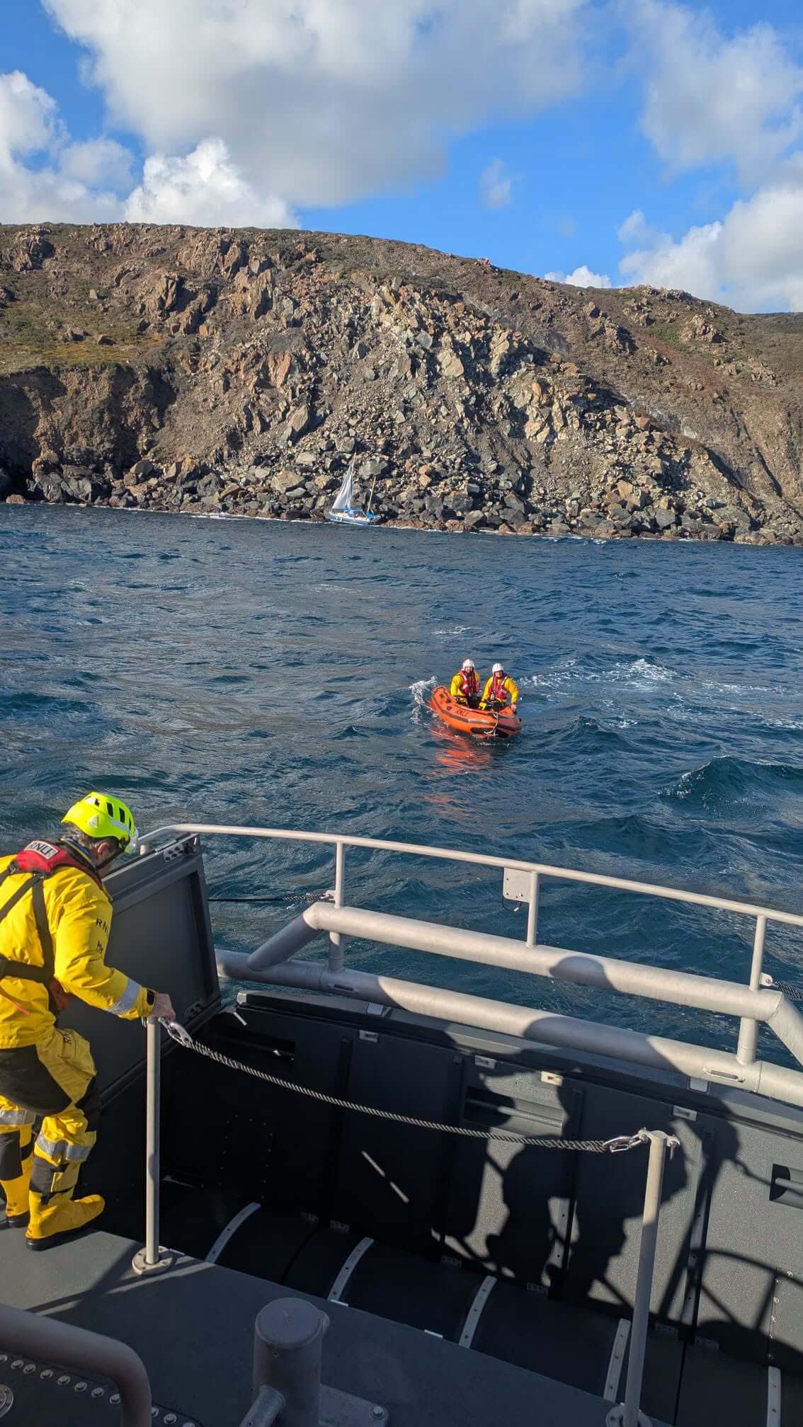 RNLI Y-boat crew in rough water near the lifeboat RNLB Rose, with the yacht on the rocks behind