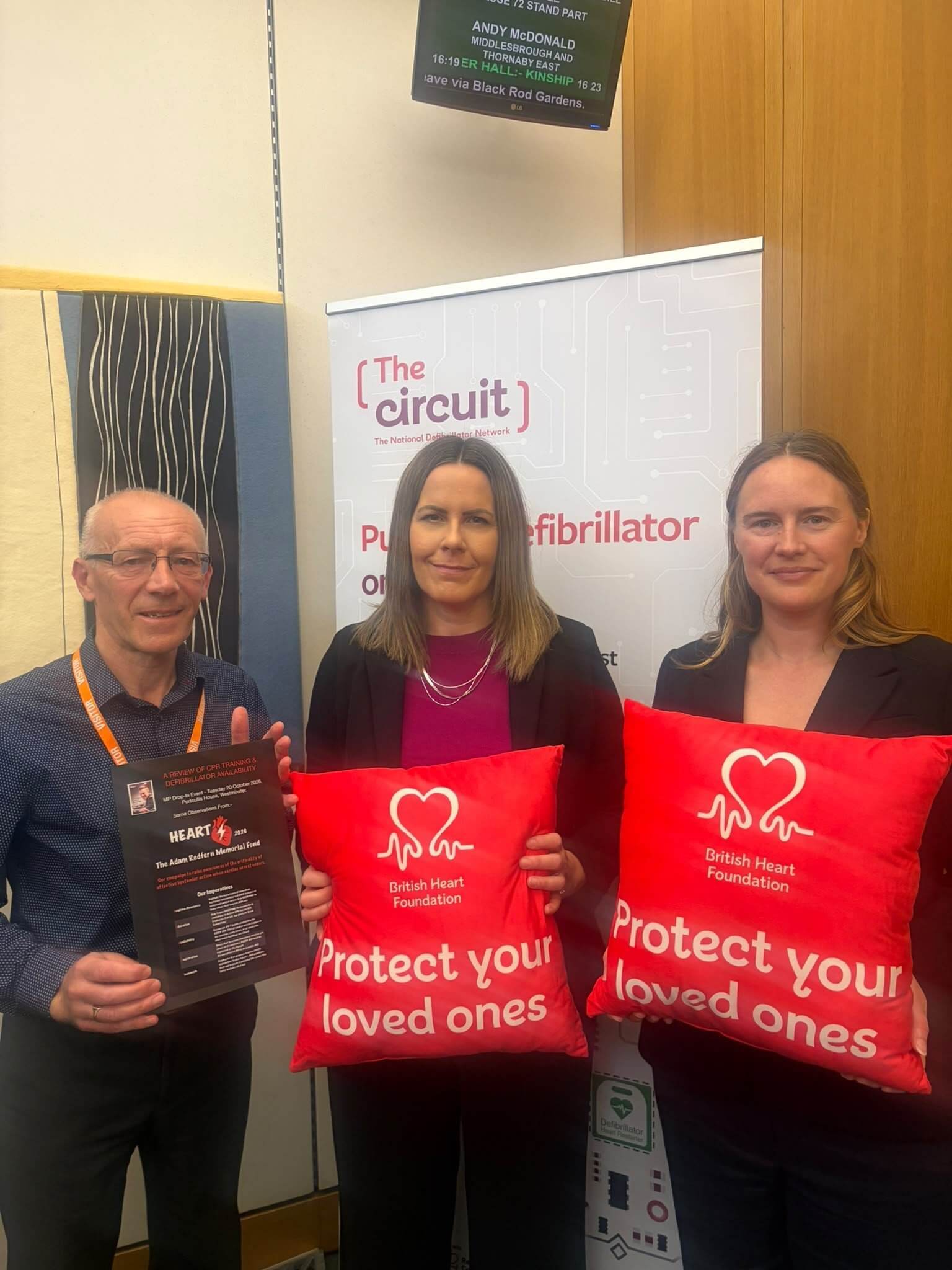 Three people in Parliament holding British Heart Foundation cushions and a defibrillator campaign leaflet