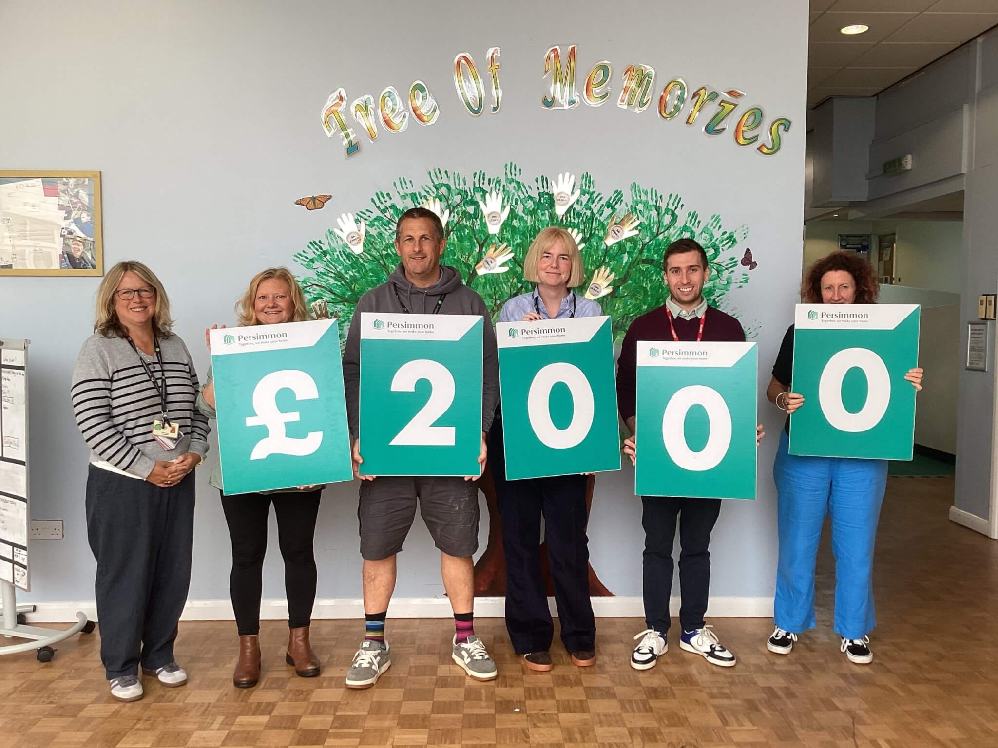 Staff and supporters holding boards spelling out £2,000 in front of the Tree of Memories mural at Doubletrees School
