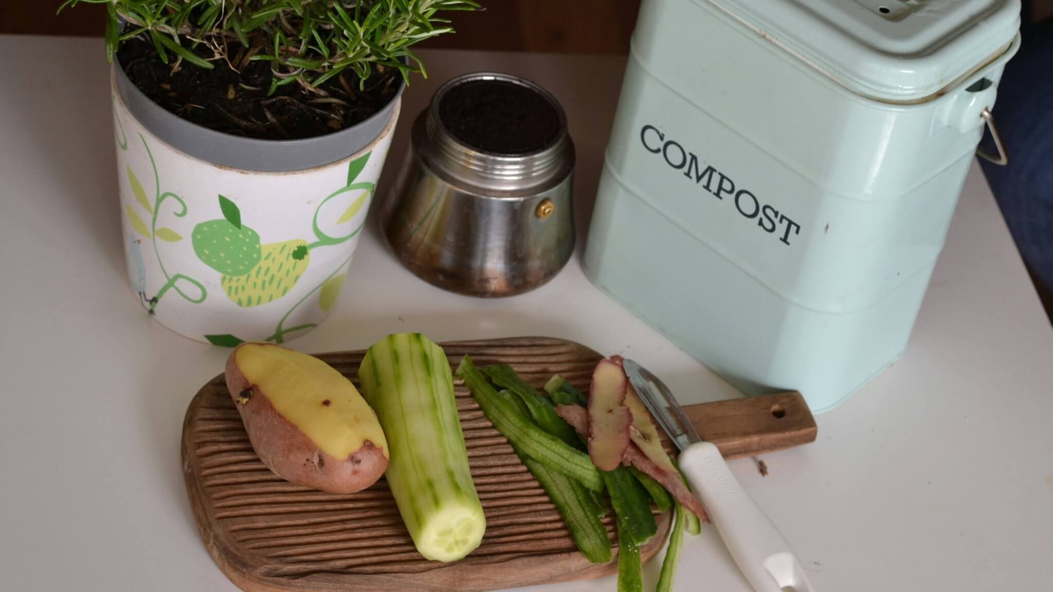 Peeled potato and cucumber with peelings on a chopping board beside a compost caddy