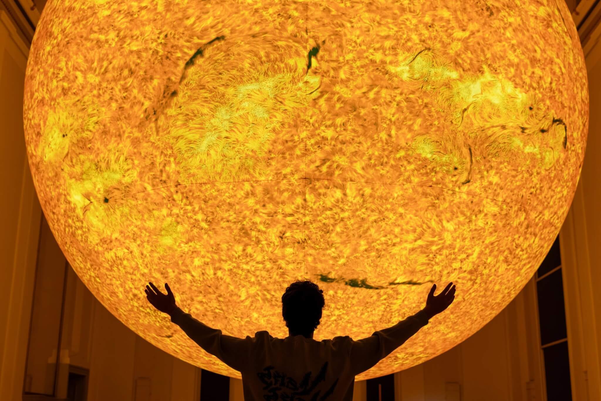 A person with arms outstretched beneath Luke Jerram's glowing Helios Sun artwork