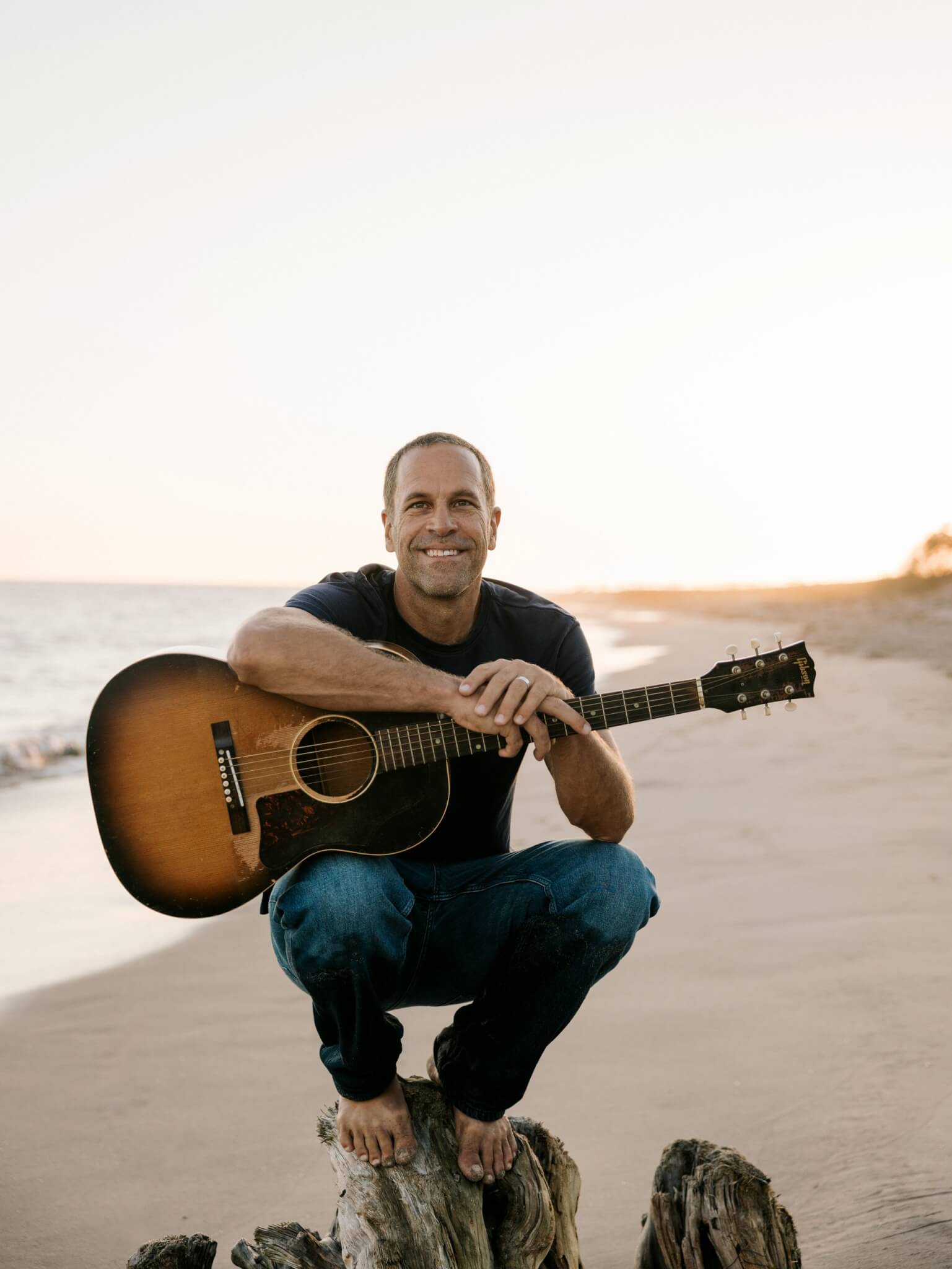 Jack Johnson crouching on a beach with his guitar