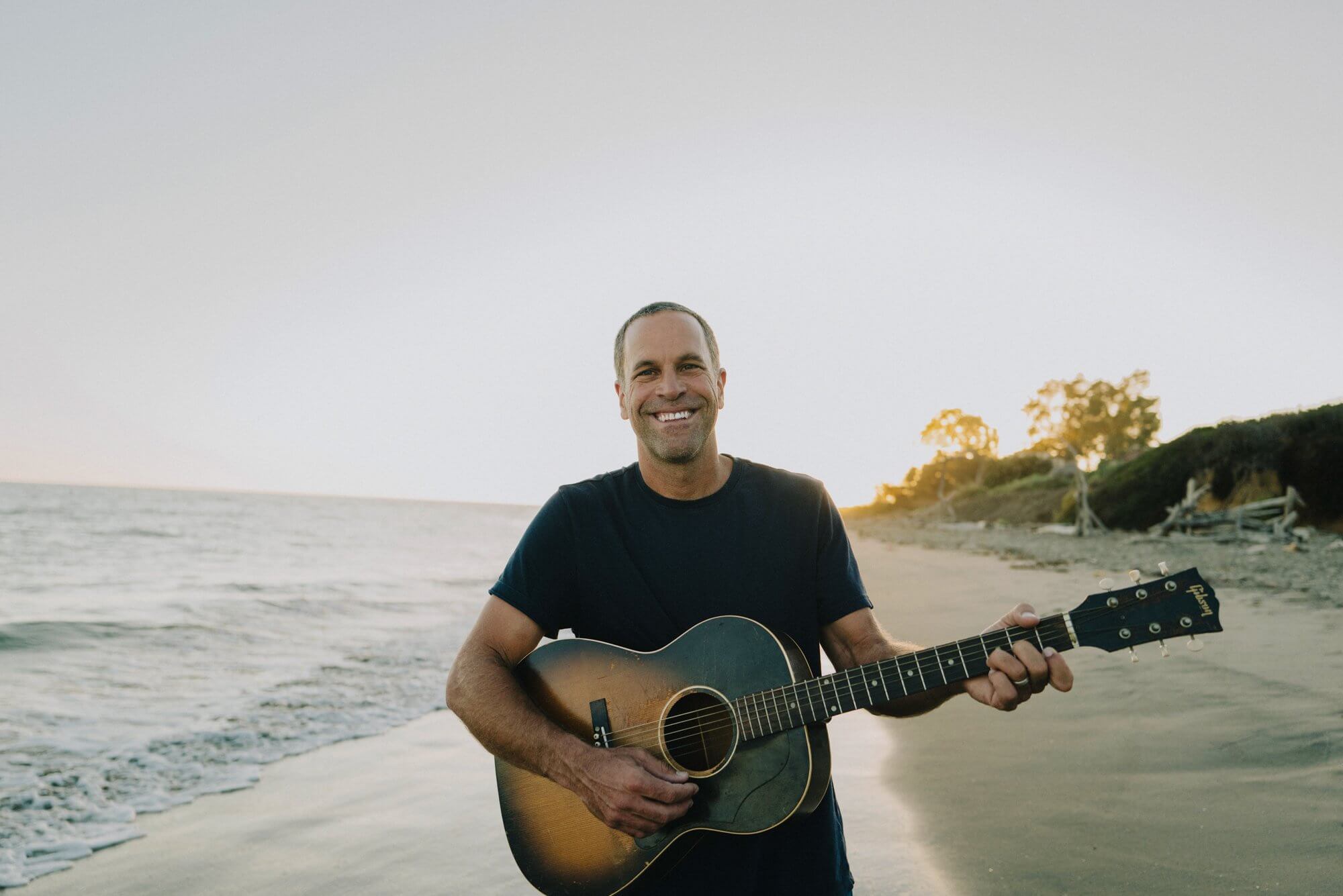 Jack Johnson holding an acoustic guitar on a beach