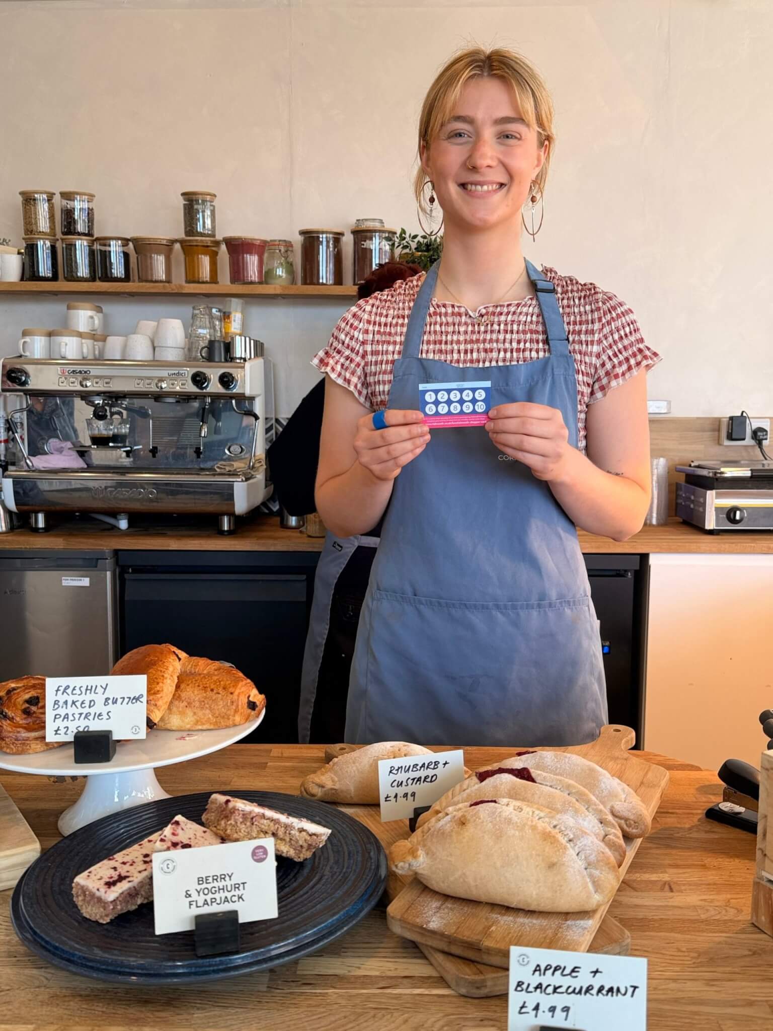 A member of staff at Oggies holding a LoveFalmouth Shopper Card behind a counter of pasties and pastries