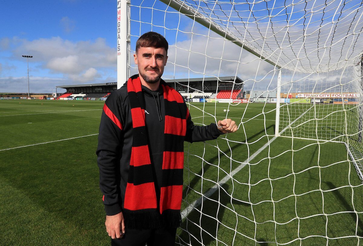 Will Dean in a red and black Truro City scarf beside a goal at Truro City Stadium