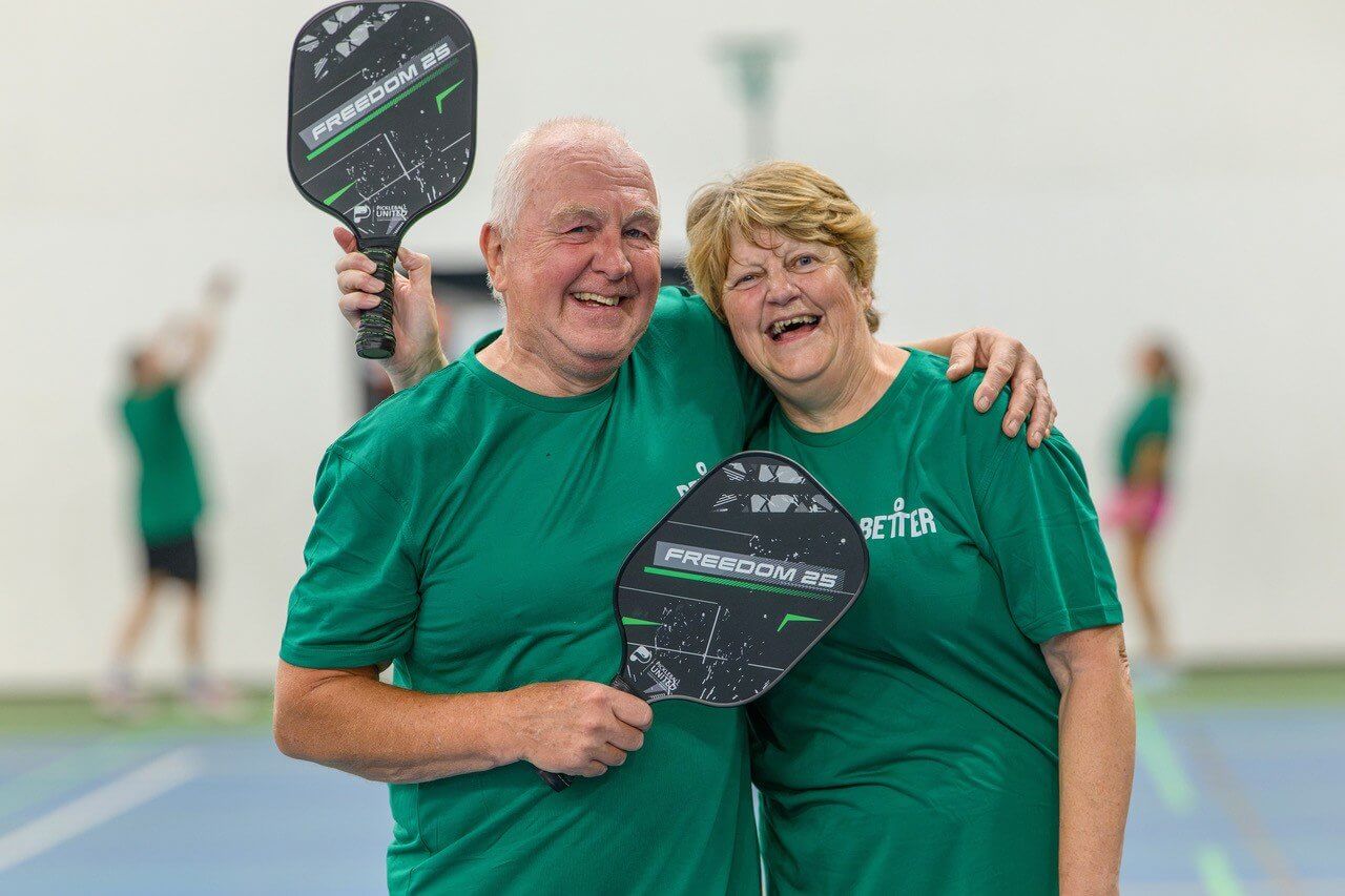 Two pickleball players in green shirts laughing with their paddles