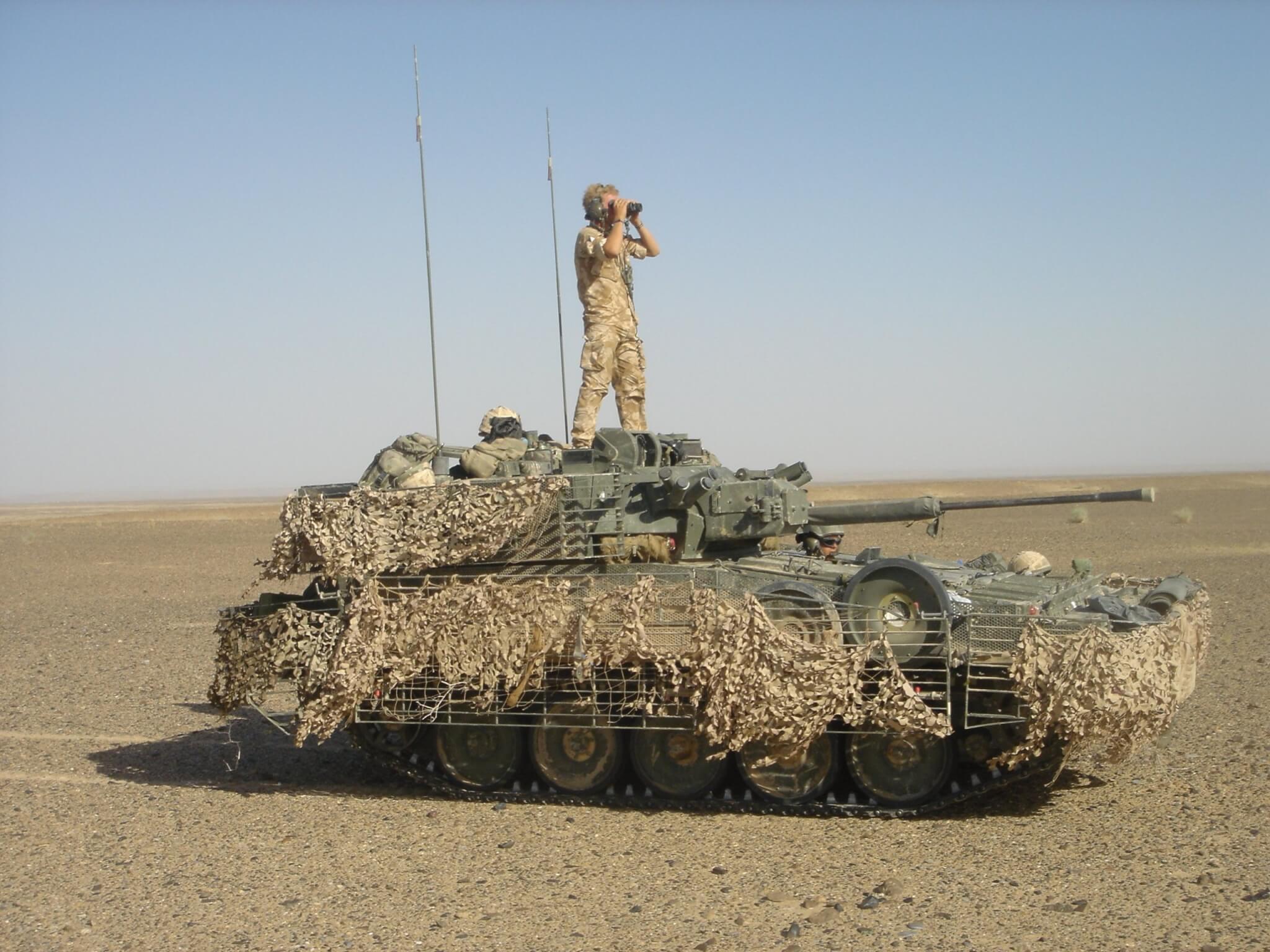 A Scimitar Mk II light tank draped in camouflage netting with a soldier standing on top looking through binoculars