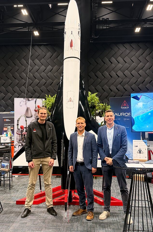 Three men standing in front of a model of Dawn Aerospace's Aurora spaceplane at an exhibition stand