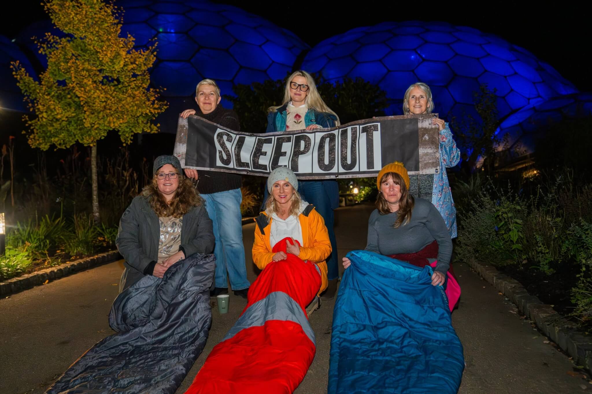 Sleep Out participants in sleeping bags holding a Sleep Out banner in front of the lit-up Eden Project Biomes