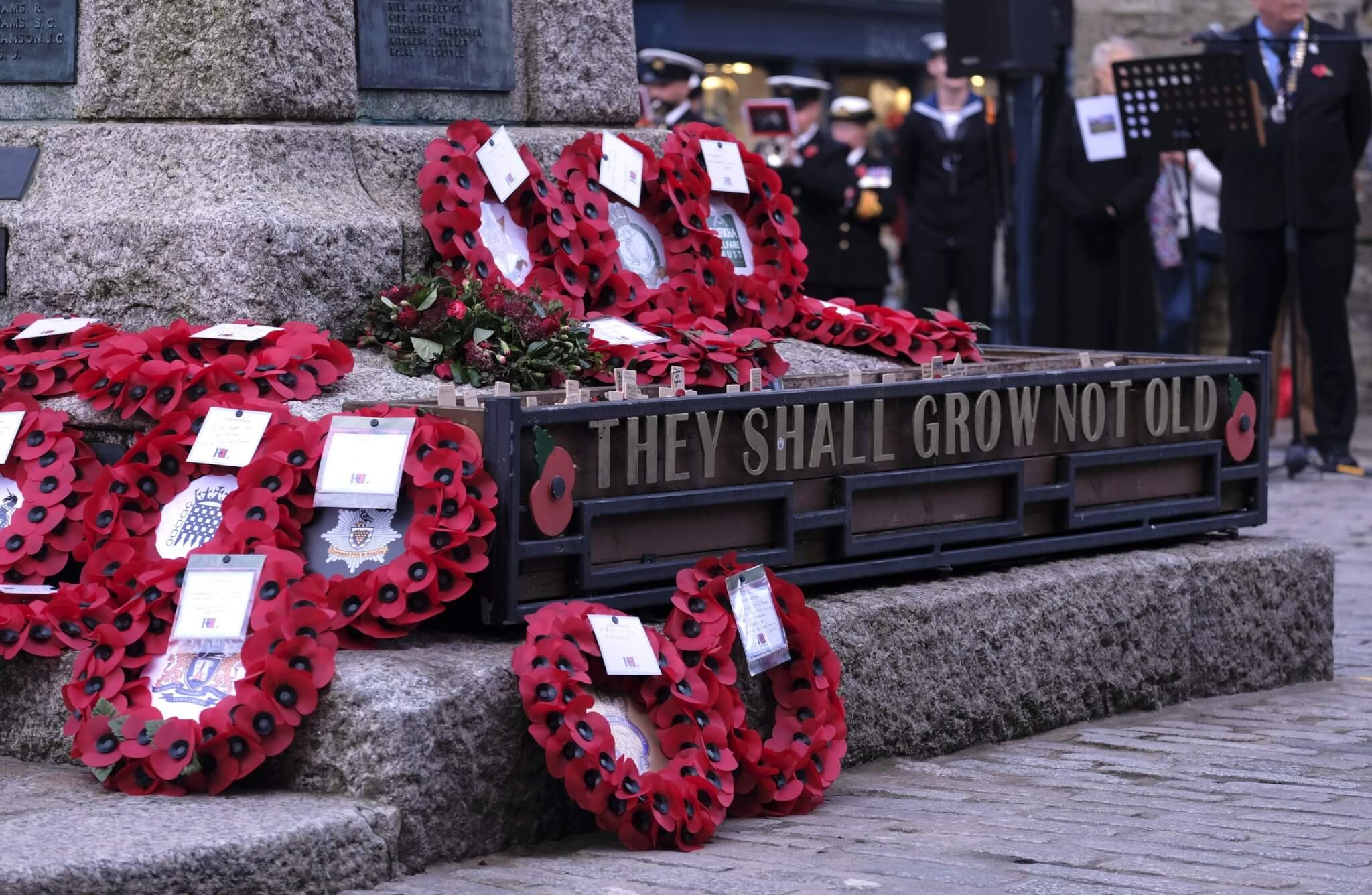 Poppy wreaths laid at the Truro War Memorial beside a planter reading They Shall Grow Not Old