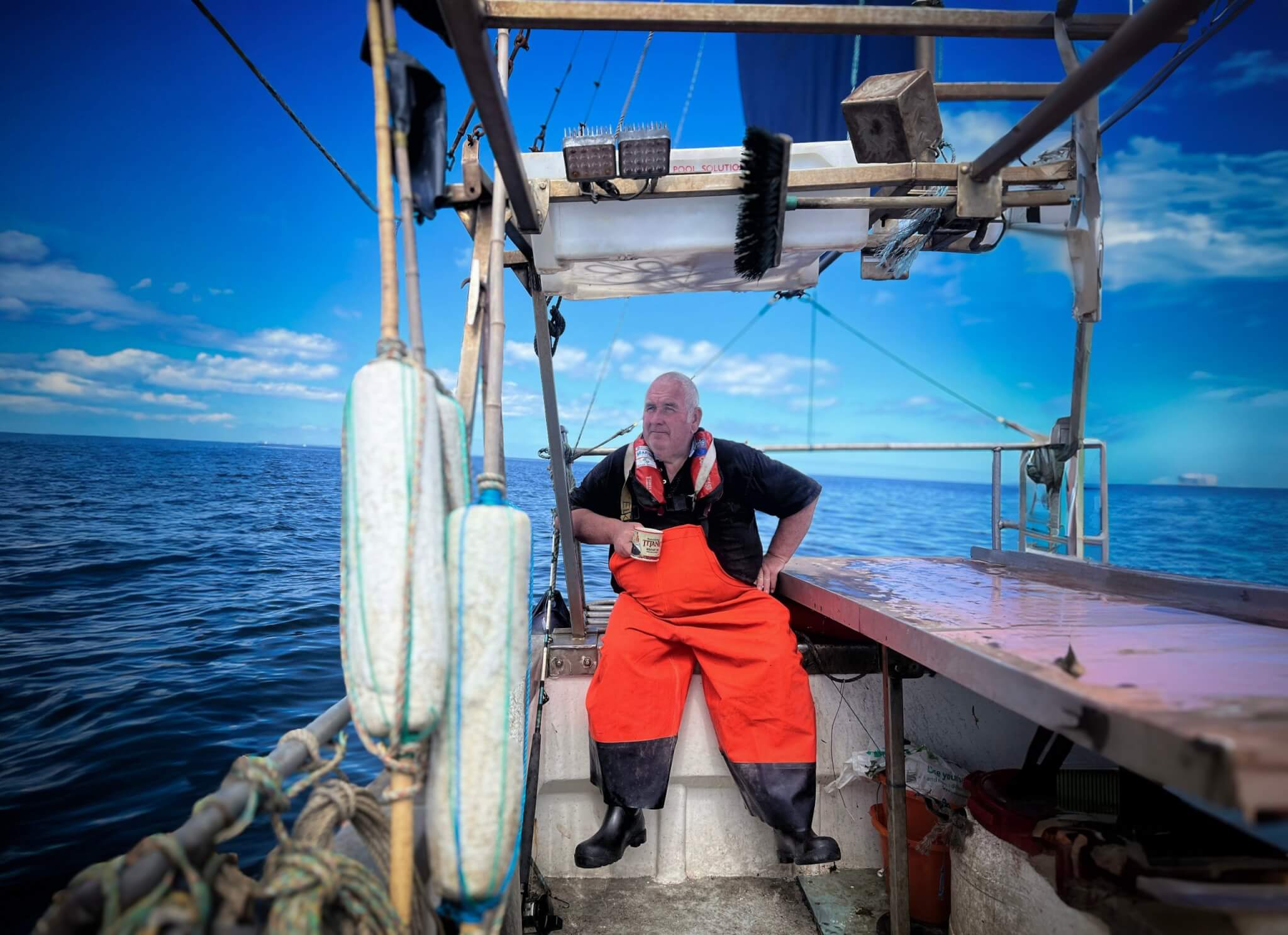 Fisherman in orange waterproofs on a fishing boat at sea, from All Rivers Spill Their Stories to the Sea