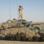 A Scimitar Mk II light tank draped in camouflage netting with a soldier standing on top looking through binoculars