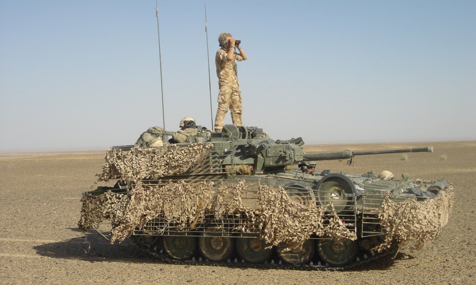 A Scimitar Mk II light tank draped in camouflage netting with a soldier standing on top looking through binoculars