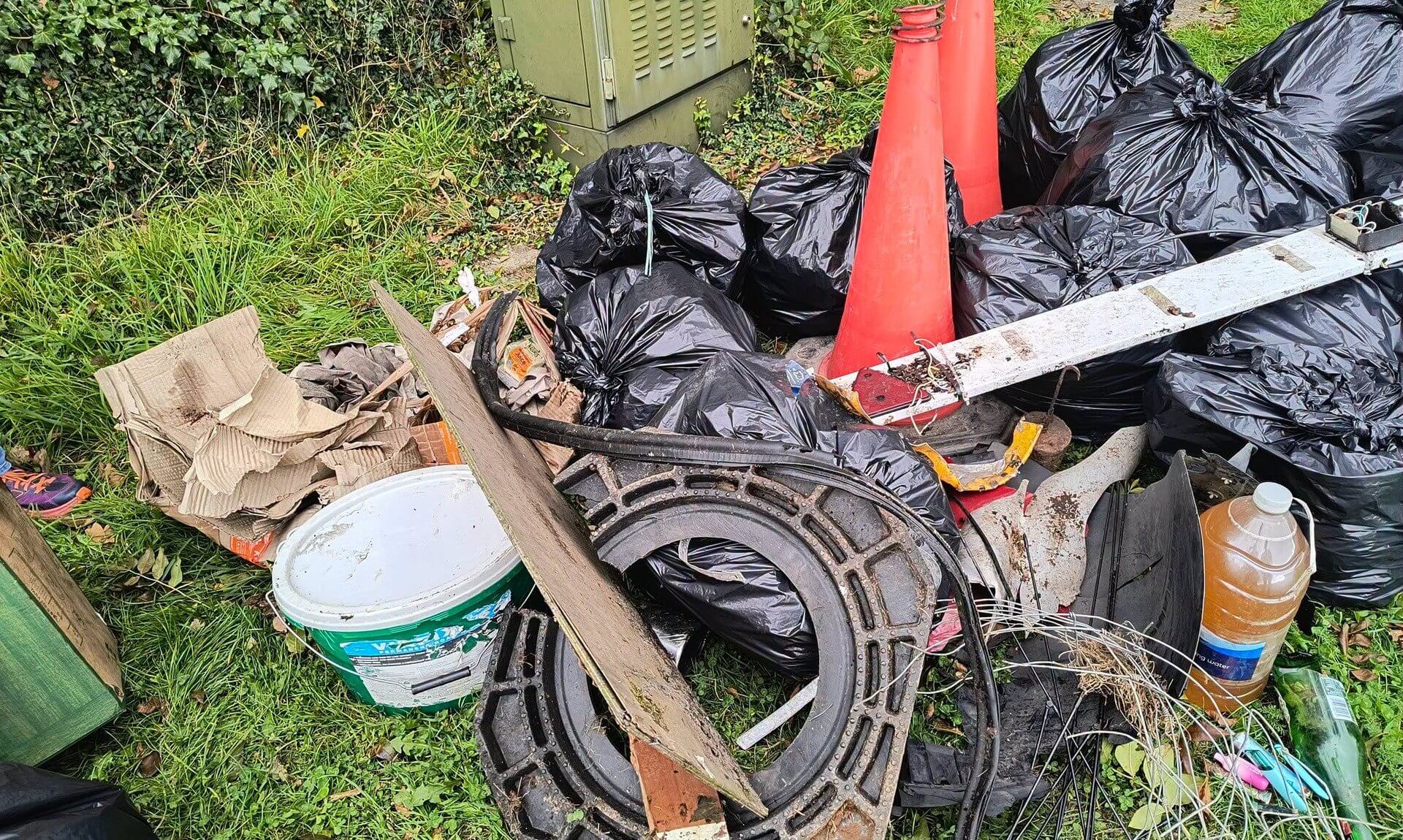 Black bin bags, two traffic cones, a bucket, cardboard and wire piled on a grass verge in Bodmin