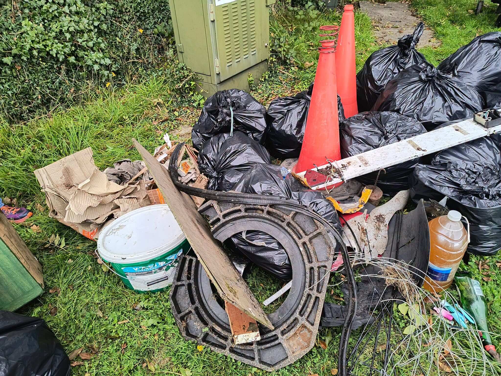 Black bin bags, two traffic cones, a bucket, cardboard and wire piled on a grass verge in Bodmin