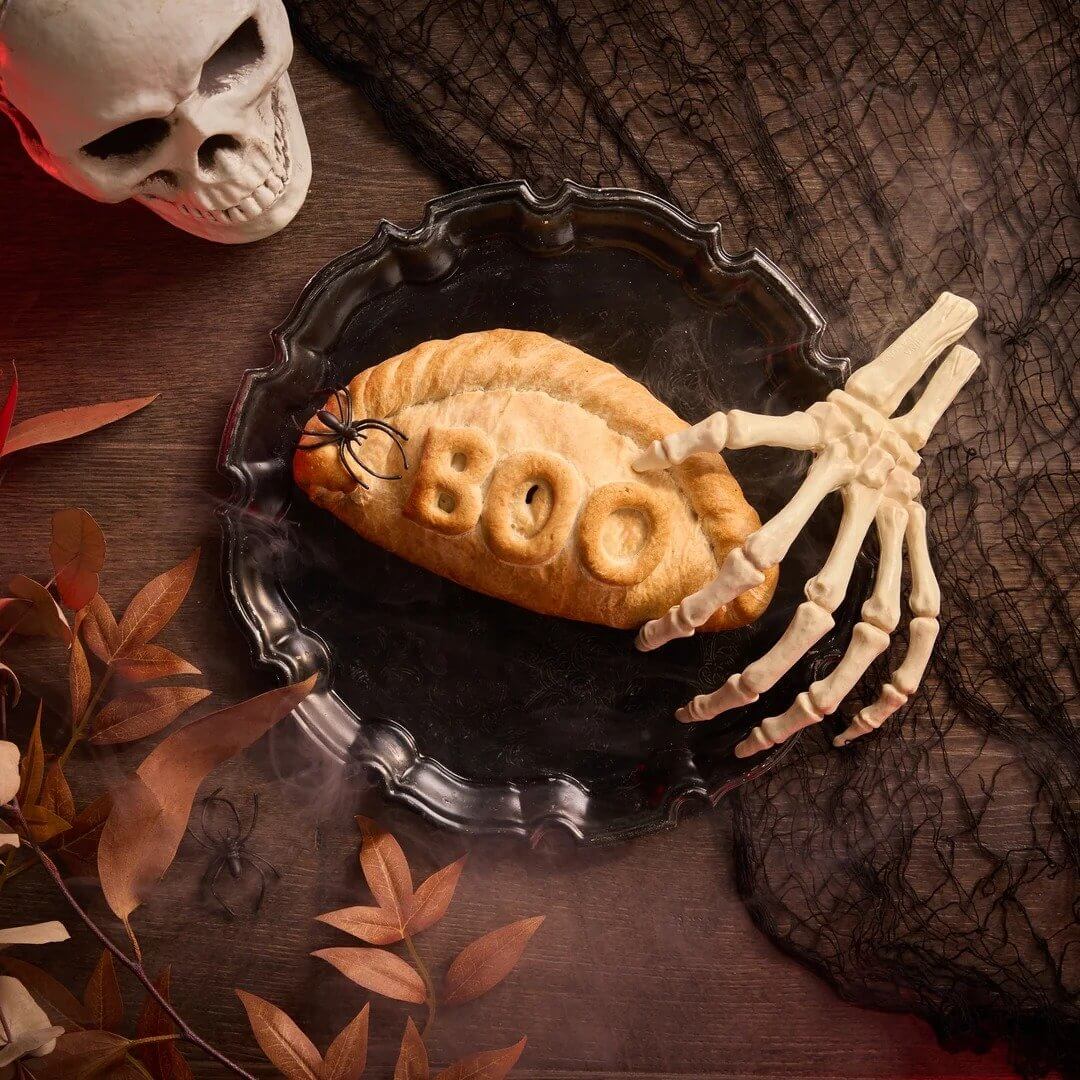 Cornish steak pasty stamped with BOO on a black tray beside a skeleton hand