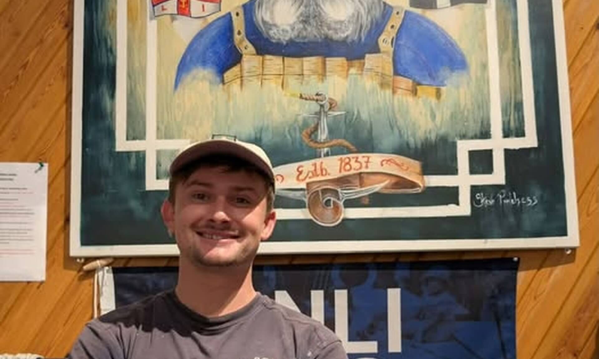 Bude RNLI crew member Noah Lakeland in front of a lifeboat painting at the station