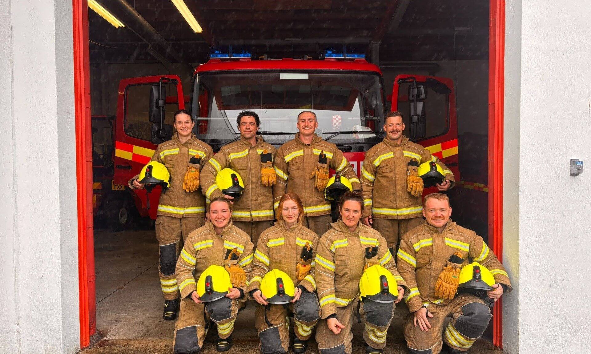 Eight firefighters in kit in front of a fire engine at a fire station