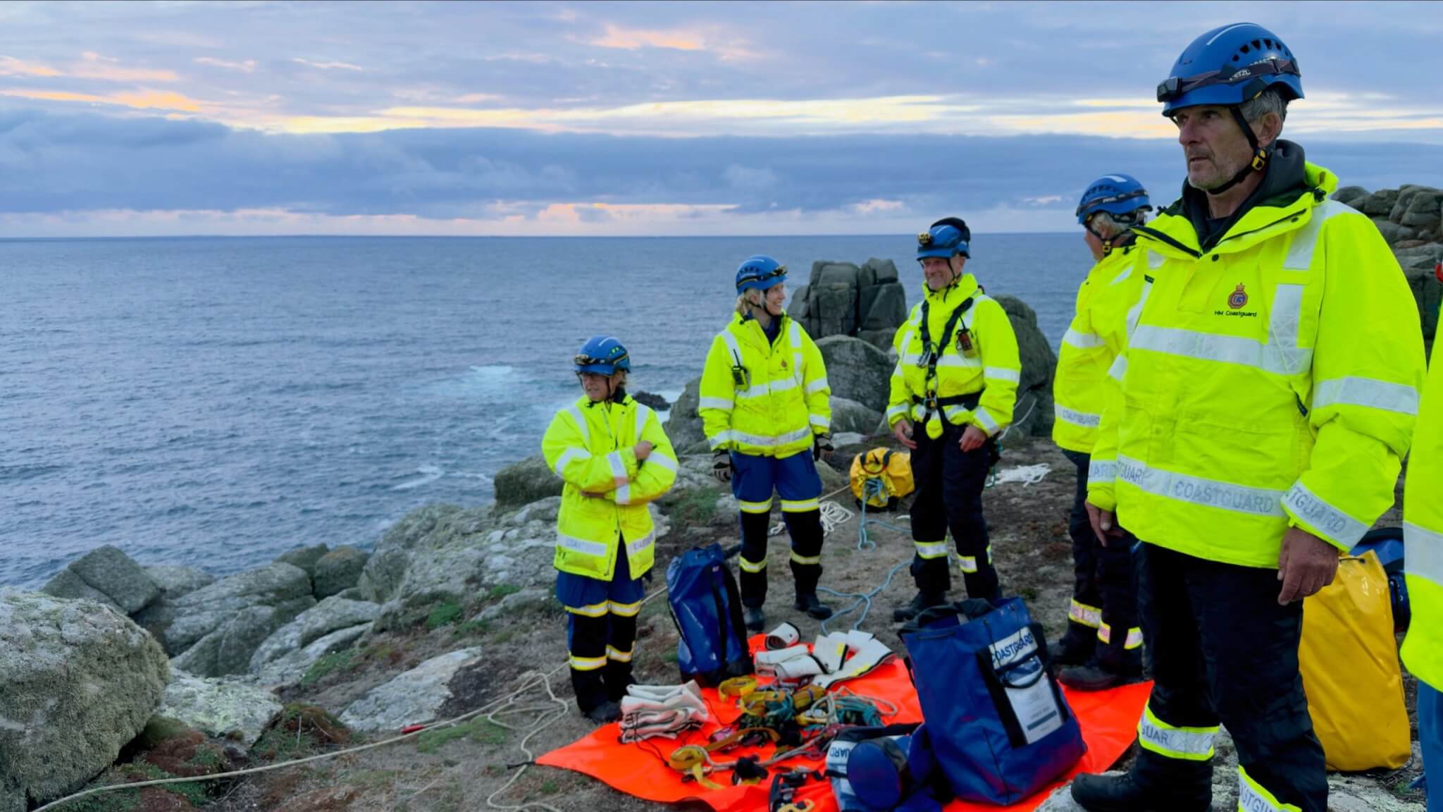 Coastguard officers in high-vis and helmets with rope rescue gear on a cliff above the sea
