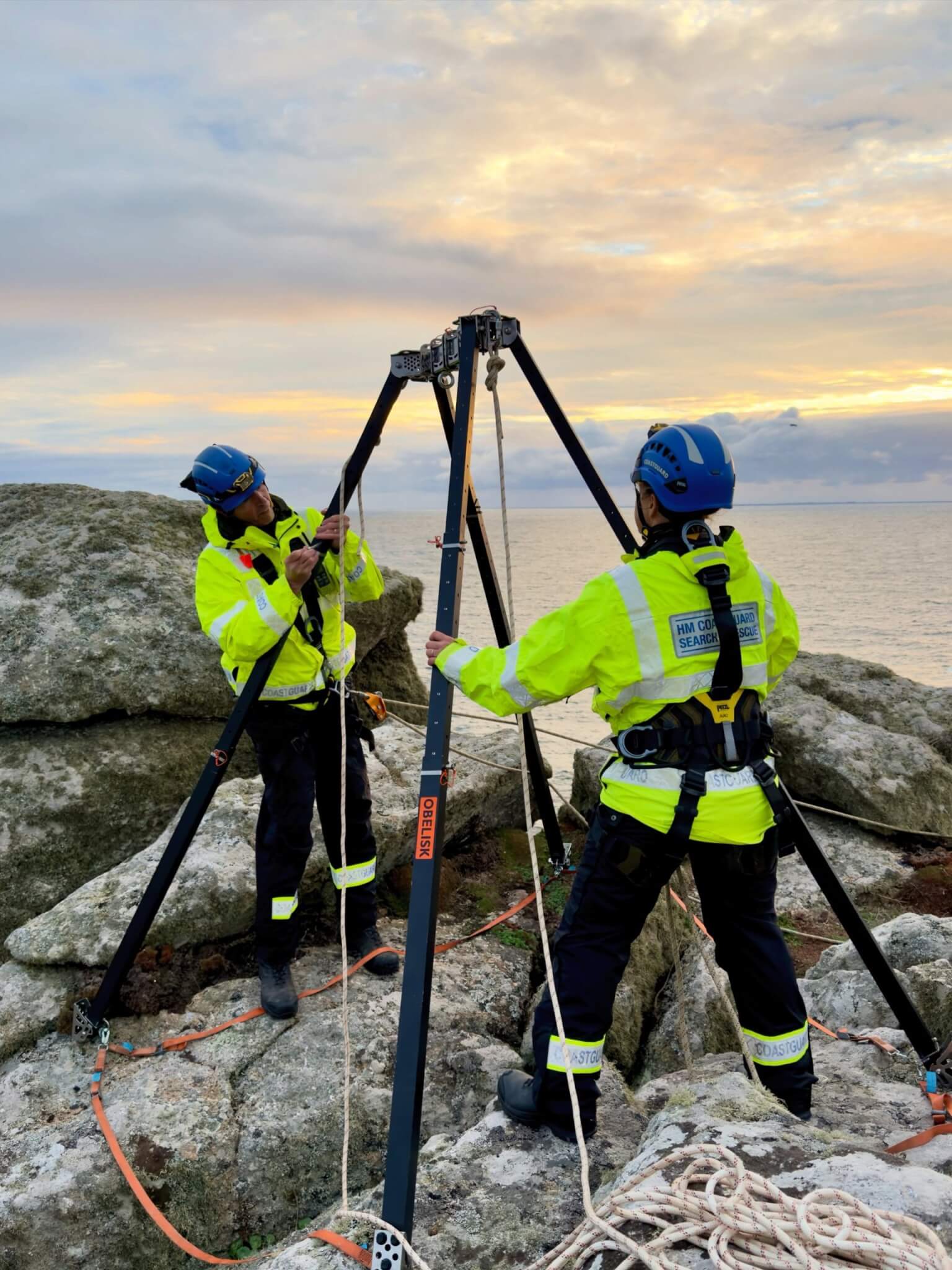 Two coastguard technicians rigging a rescue tripod at sunset