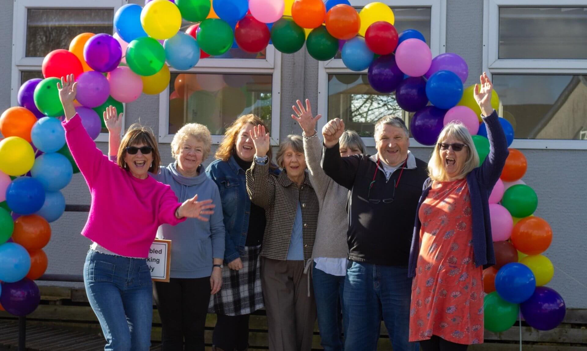 Whitemoor Village Hall committee members cheering under a rainbow balloon arch
