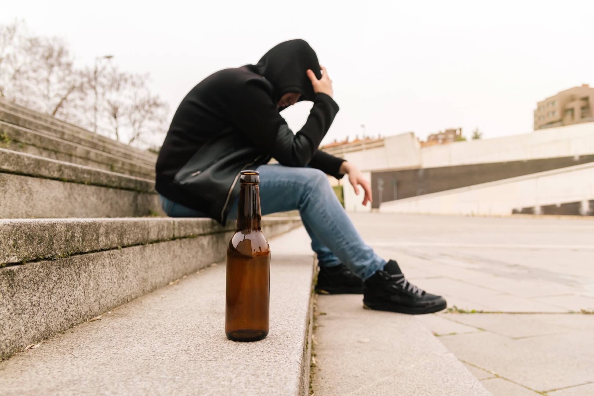 Person in a hood sitting on concrete steps with a beer bottle