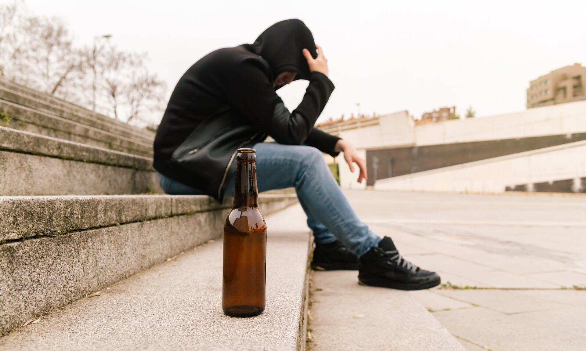 Person in a hood sitting on concrete steps with a beer bottle