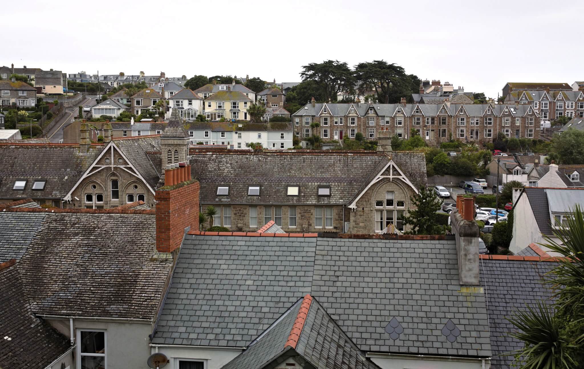 Slate rooftops and chimneys with rows of houses on the hillside behind in a Cornish town
