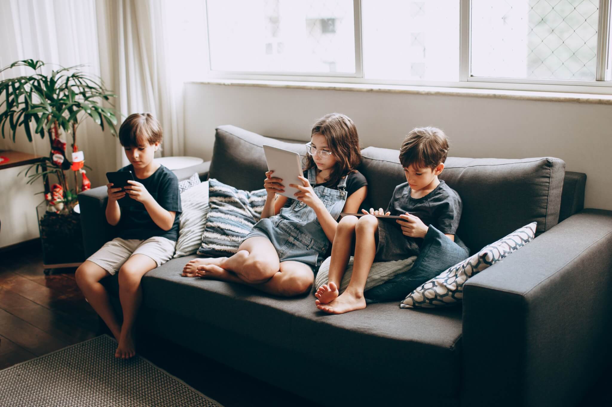 Three children sitting on a sofa, each looking at a phone or tablet