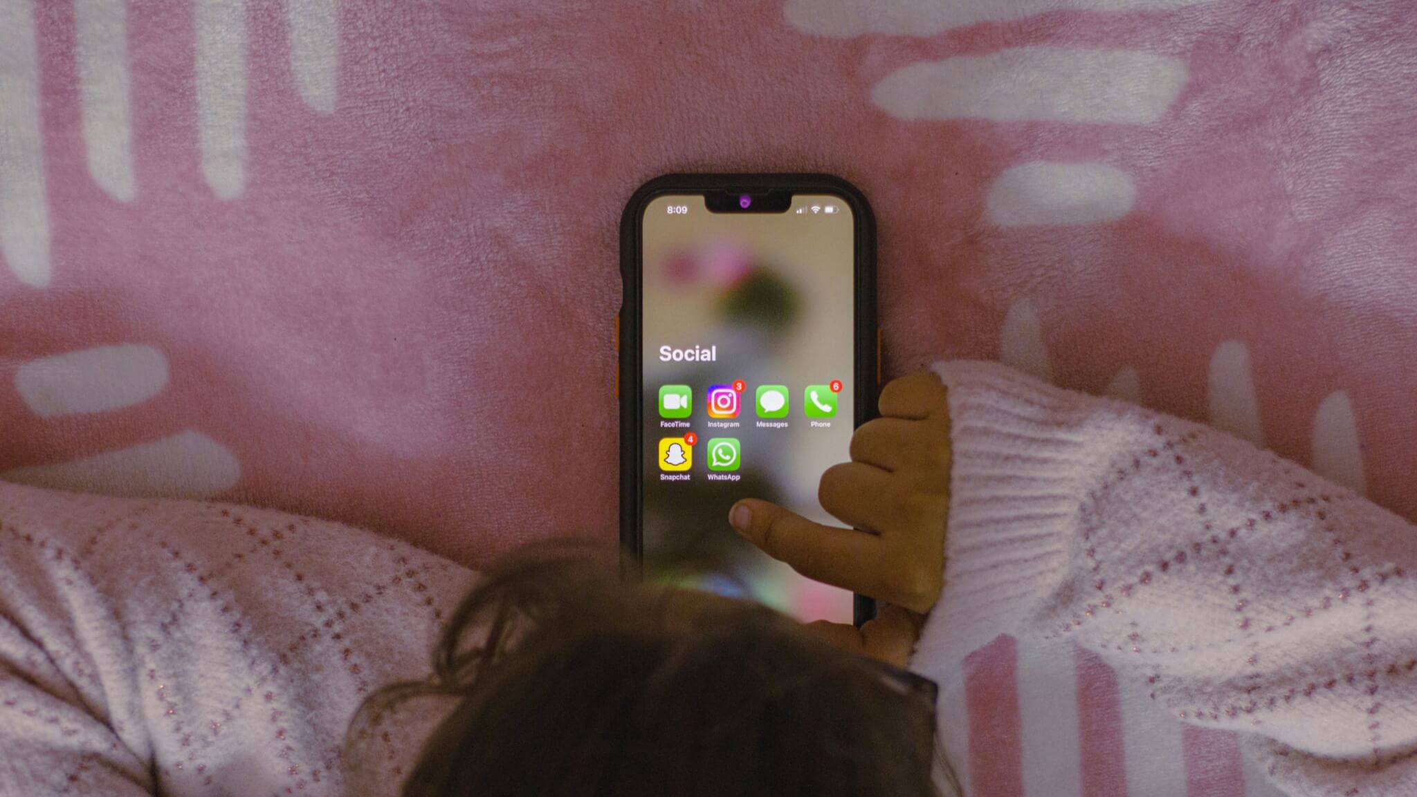 A child lying on a pink blanket looking at social media apps on a phone
