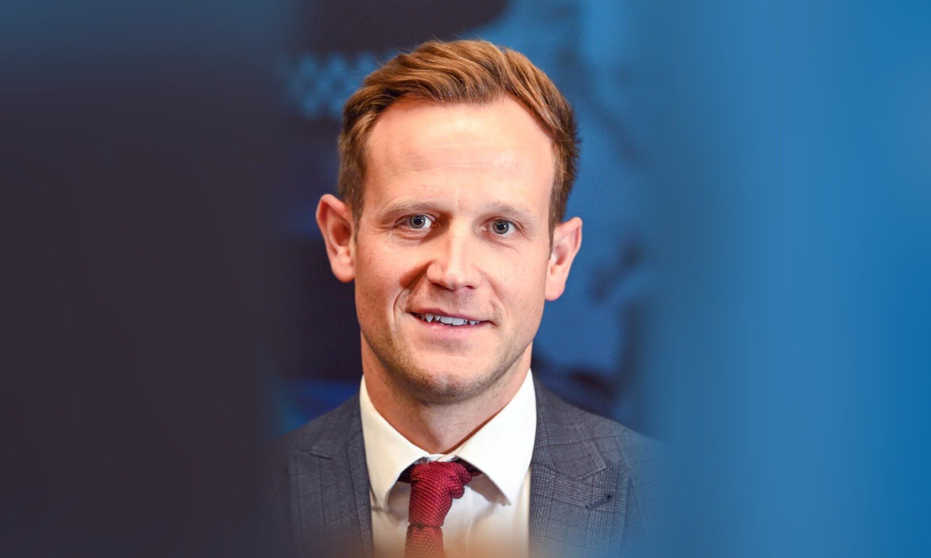 Detective in a grey checked suit and red tie in front of a blue backdrop
