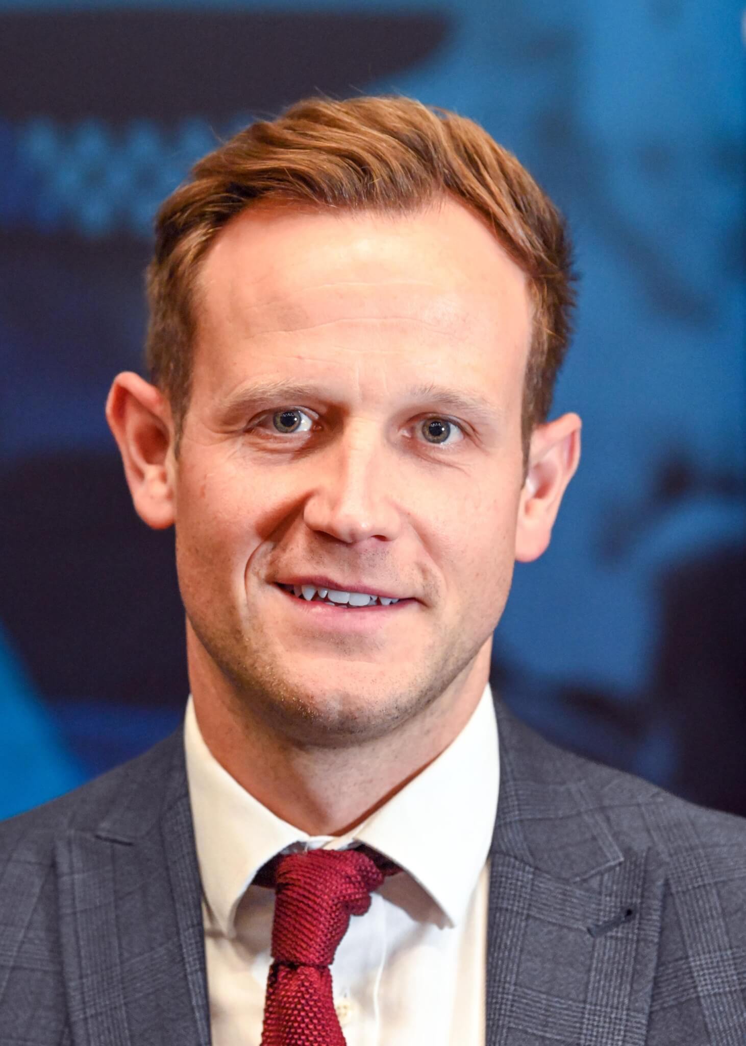 Detective in a grey checked suit and red tie in front of a blue backdrop