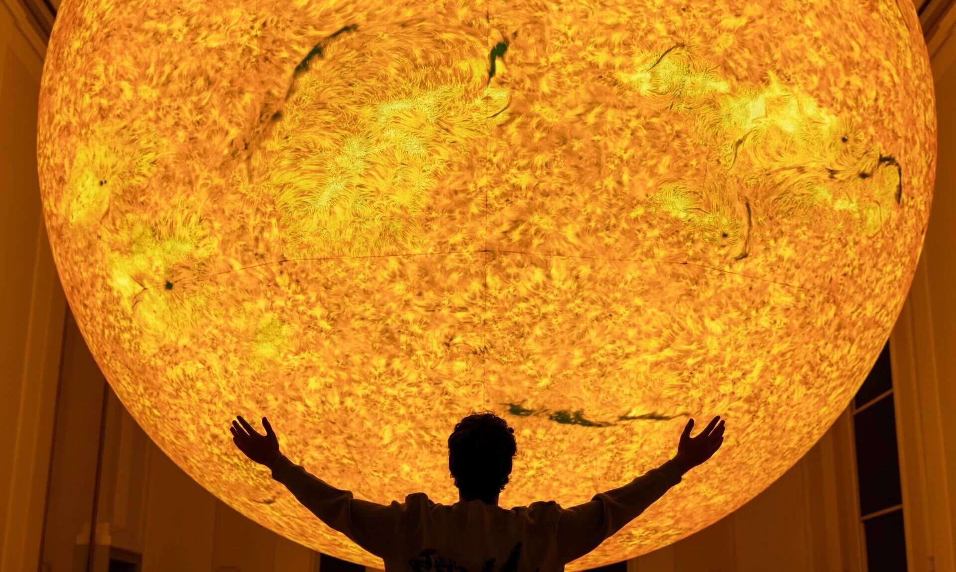 A person with arms outstretched beneath Luke Jerram's glowing Helios Sun artwork