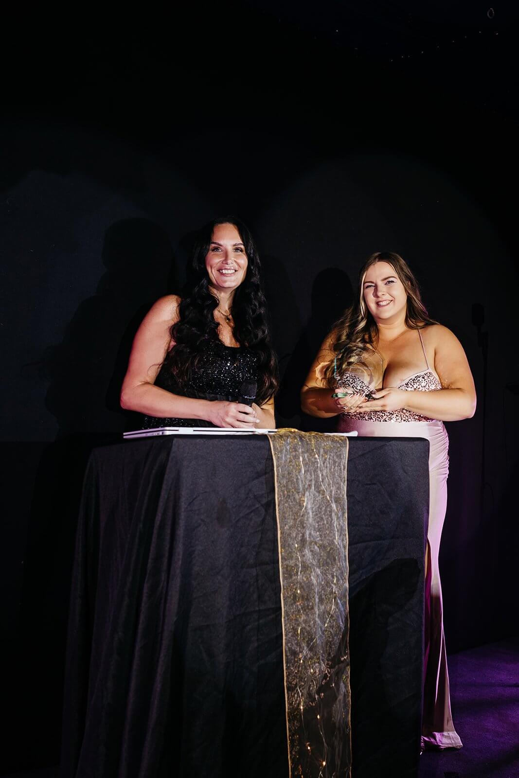Emma Chowings holding a trophy on stage beside a presenter at the Cornwall Hair and Beauty Awards