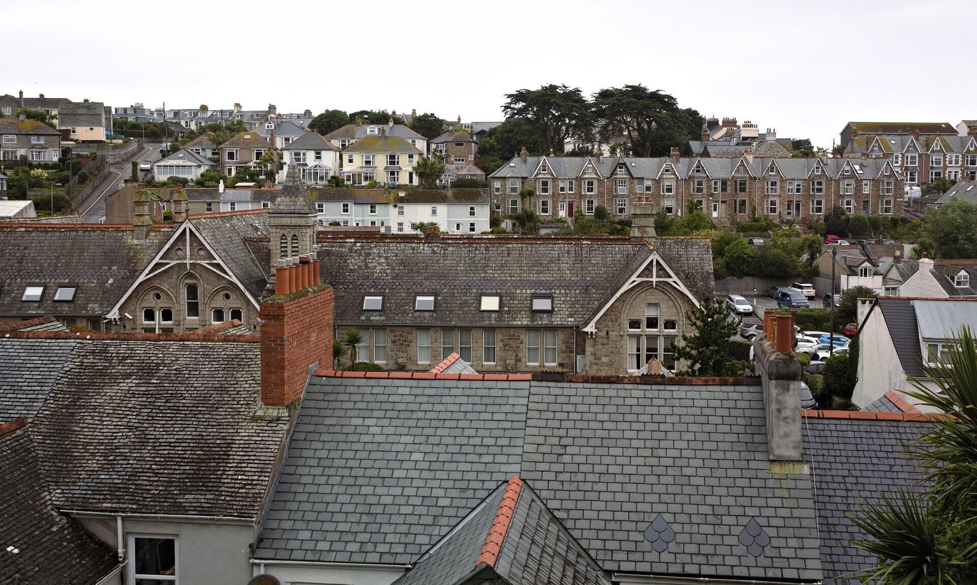 Slate rooftops and chimneys with rows of houses on the hillside behind in a Cornish town