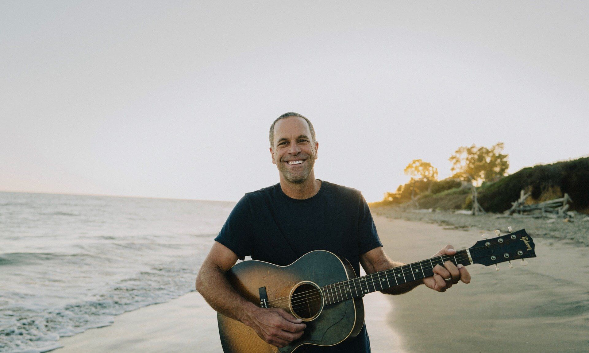Jack Johnson holding an acoustic guitar on a beach