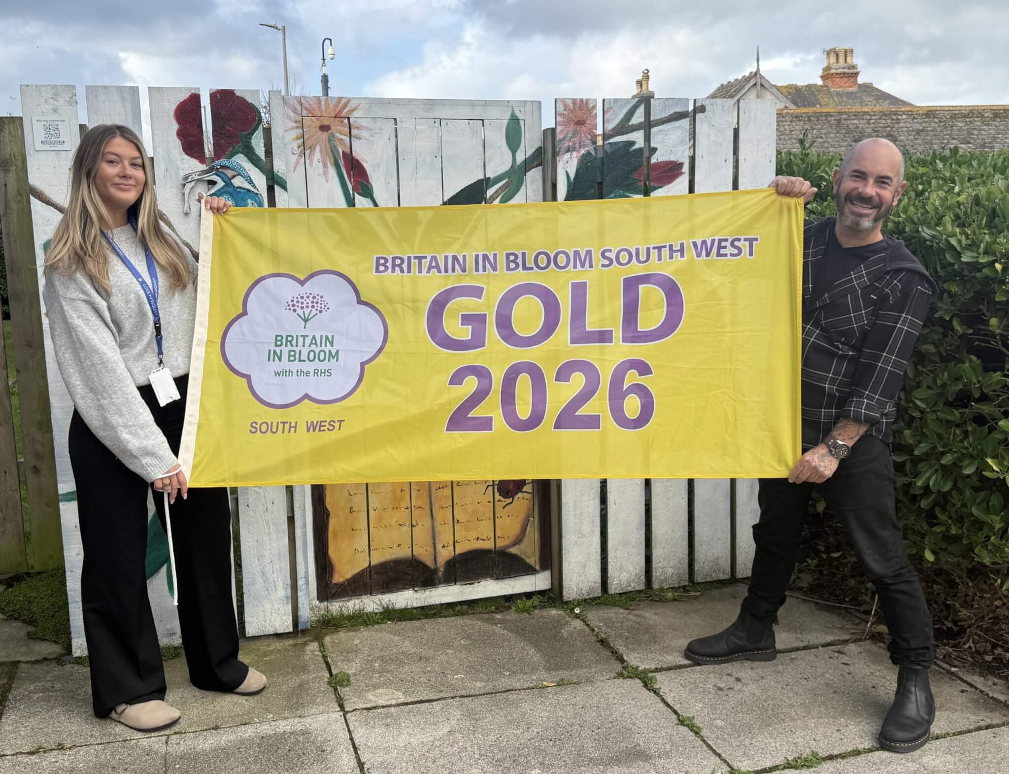 Two people holding a Britain in Bloom South West Gold 2026 flag in front of a painted fence in Newquay