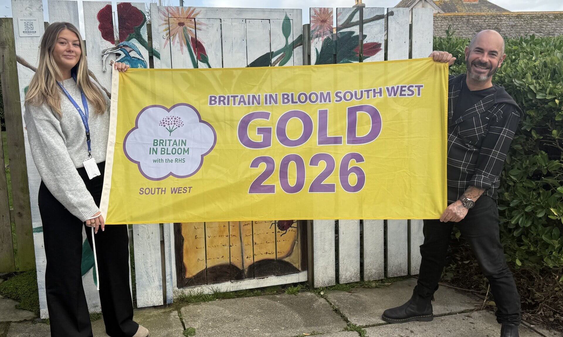Two people holding a Britain in Bloom South West Gold 2026 flag in front of a painted fence in Newquay