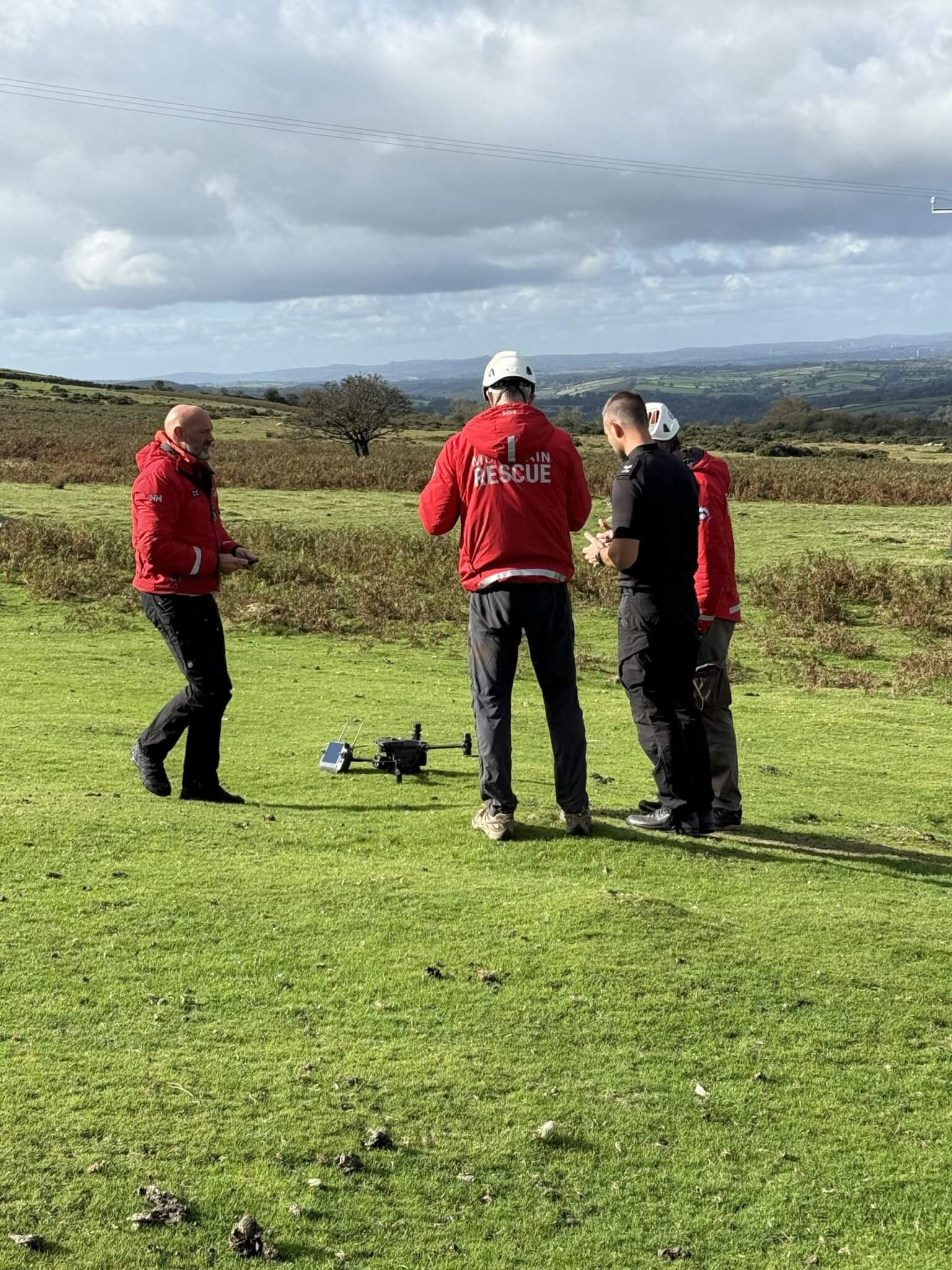 Mountain rescue volunteers and a police officer stand around a drone on open moorland