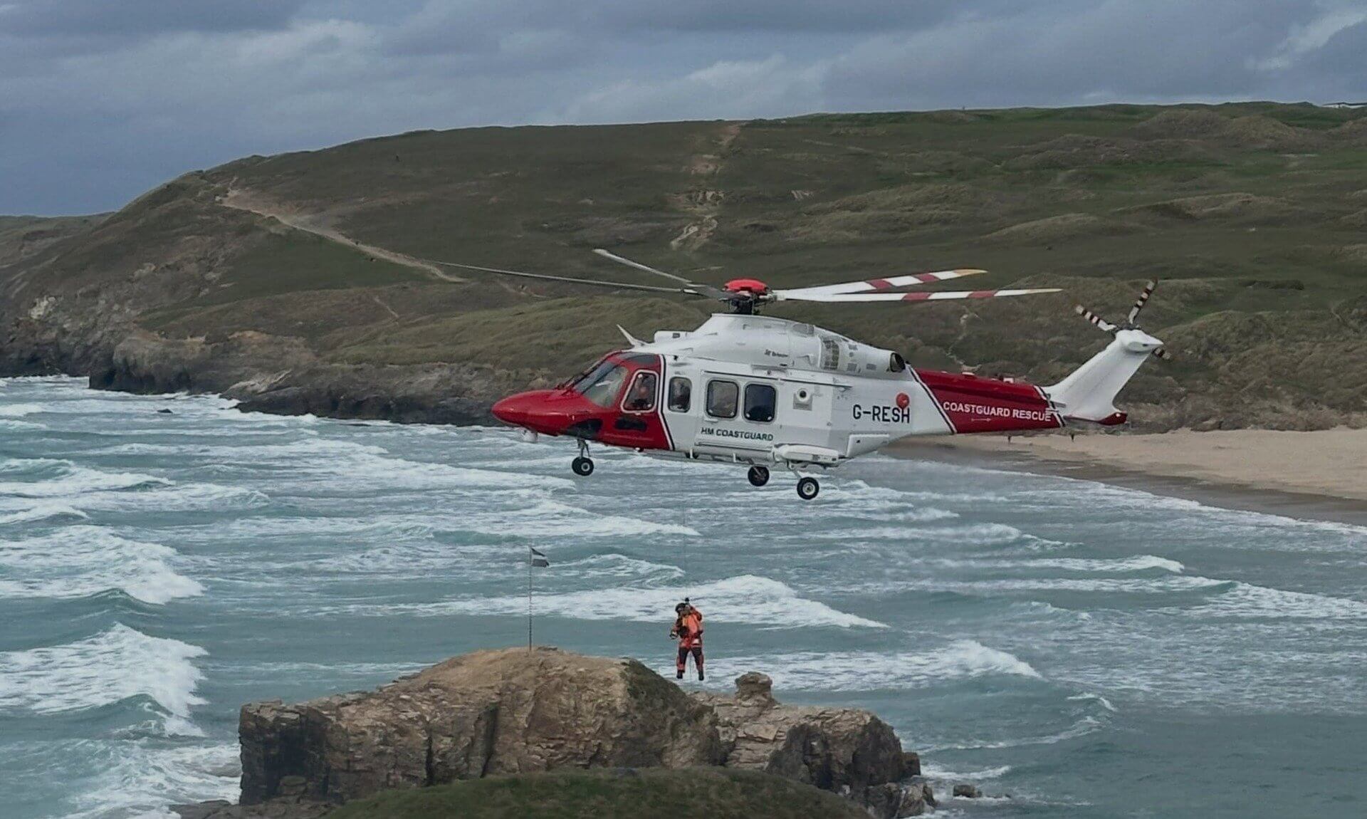Coastguard rescue helicopter hovering over a rocky outcrop at Droskyn, Perranporth, with a winchman on the wire