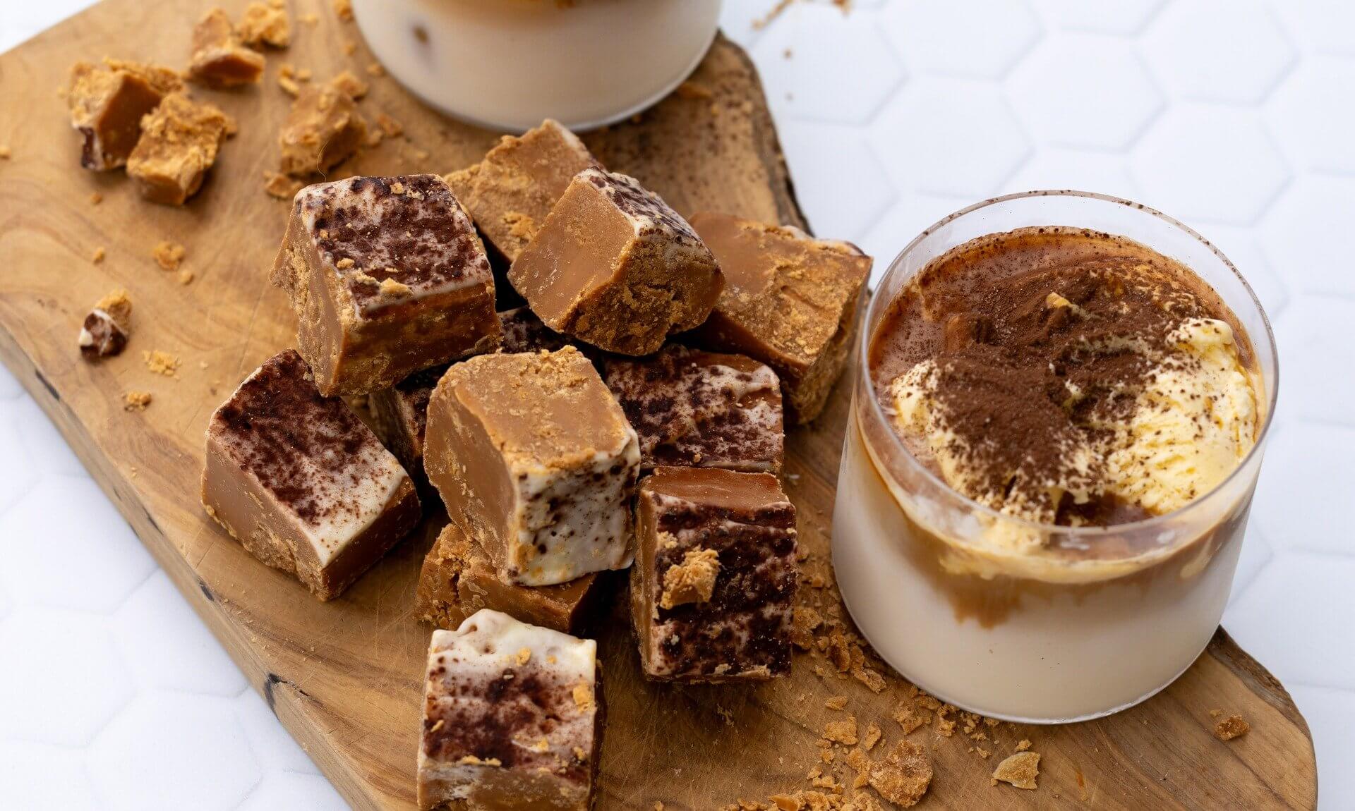 Roly's Tiramisu Fudge pieces on a wooden board beside a glass of tiramisu