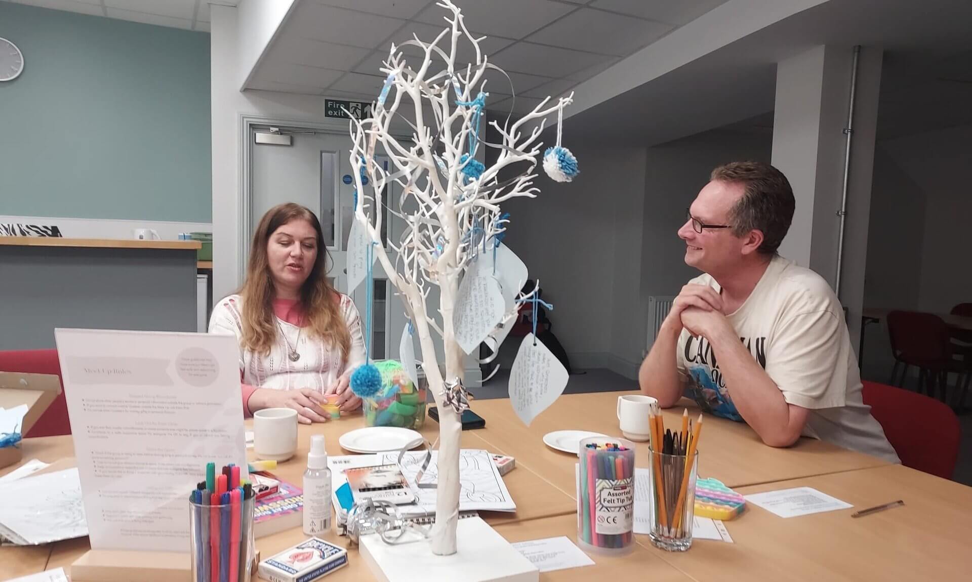 Two people chatting at a table with craft materials and a white decorative tree at a Quiet Connections Meet Up
