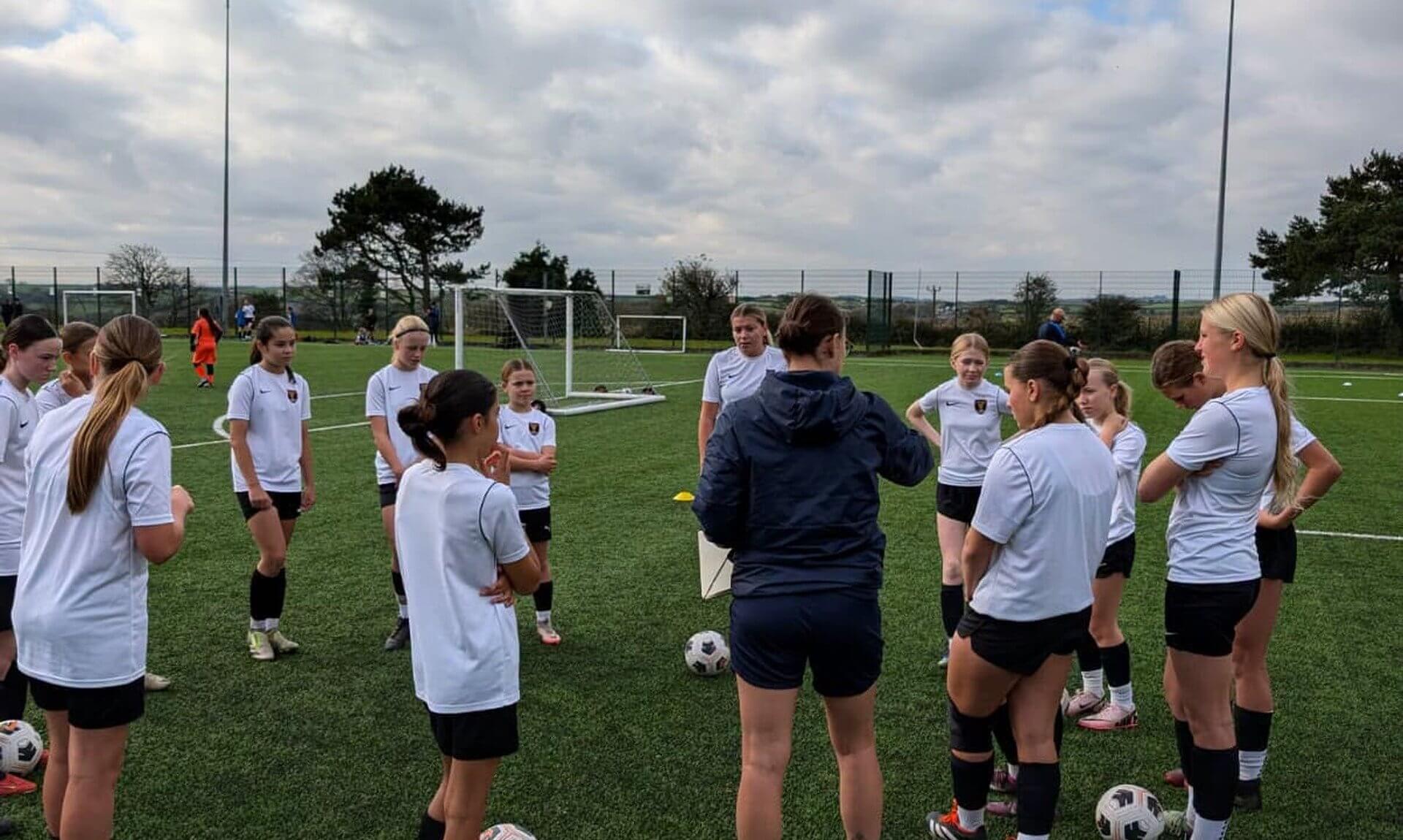 Girls in white training tops gathered around a coach on an artificial pitch