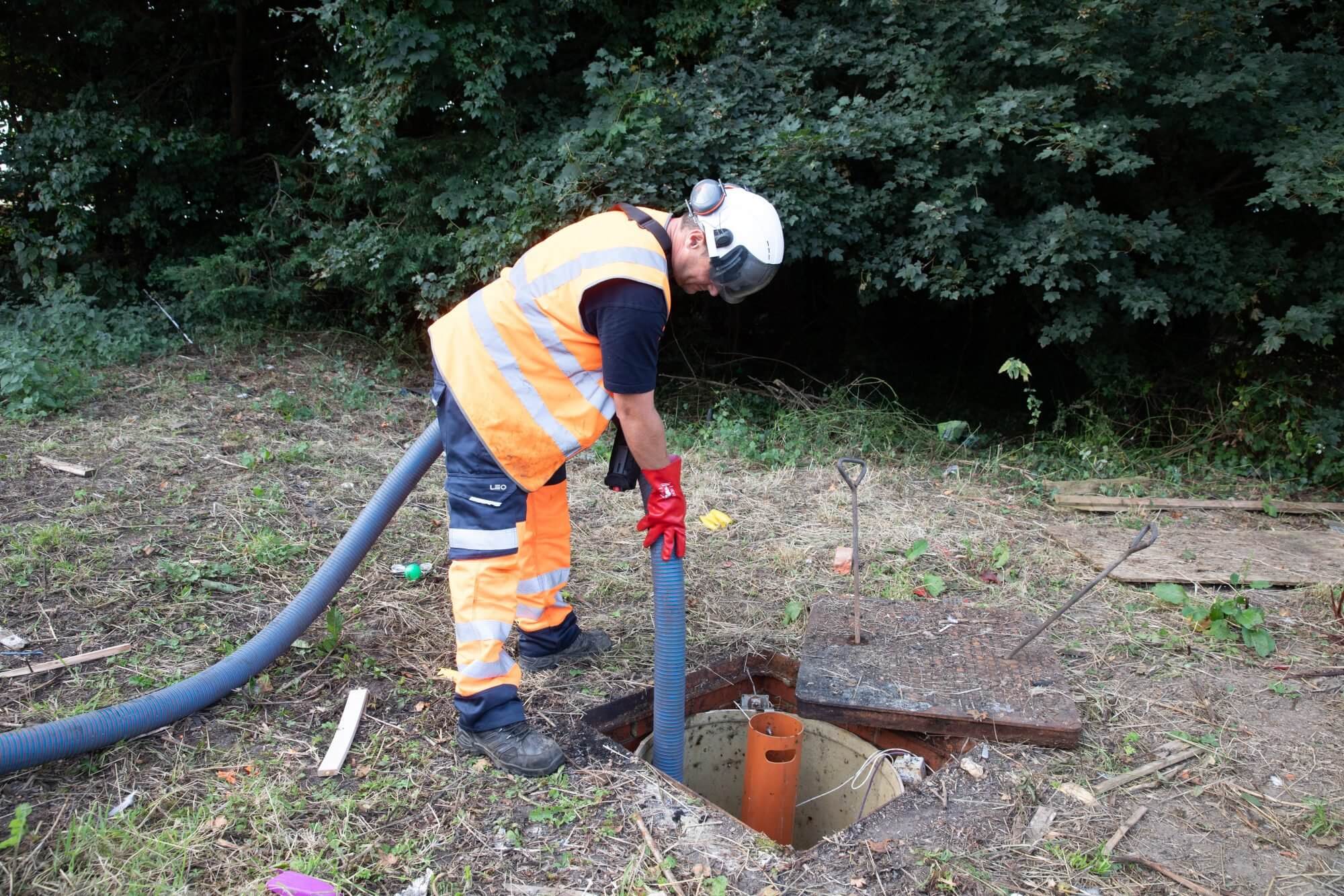 A worker in hi-vis clothing lowers a suction hose into an open septic tank
