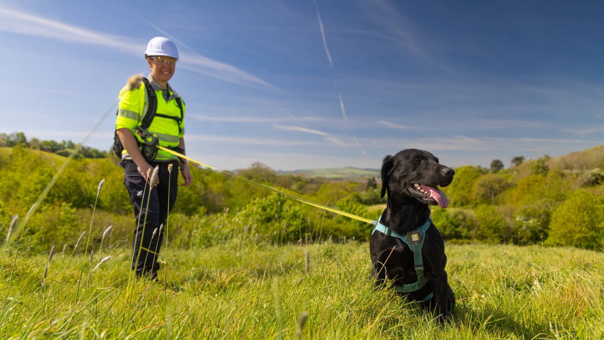 South West Water leak detection technician in hard hat and hi-vis with a black dog on a lead in a grassy field