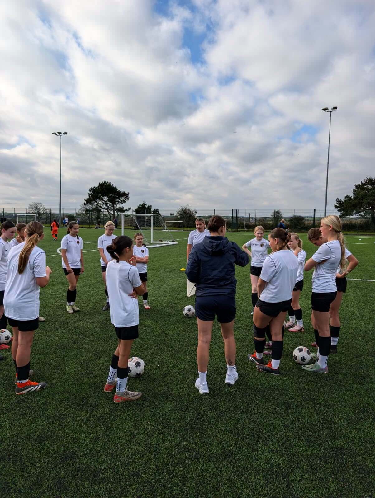 Girls in white training tops gathered around a coach on an artificial pitch