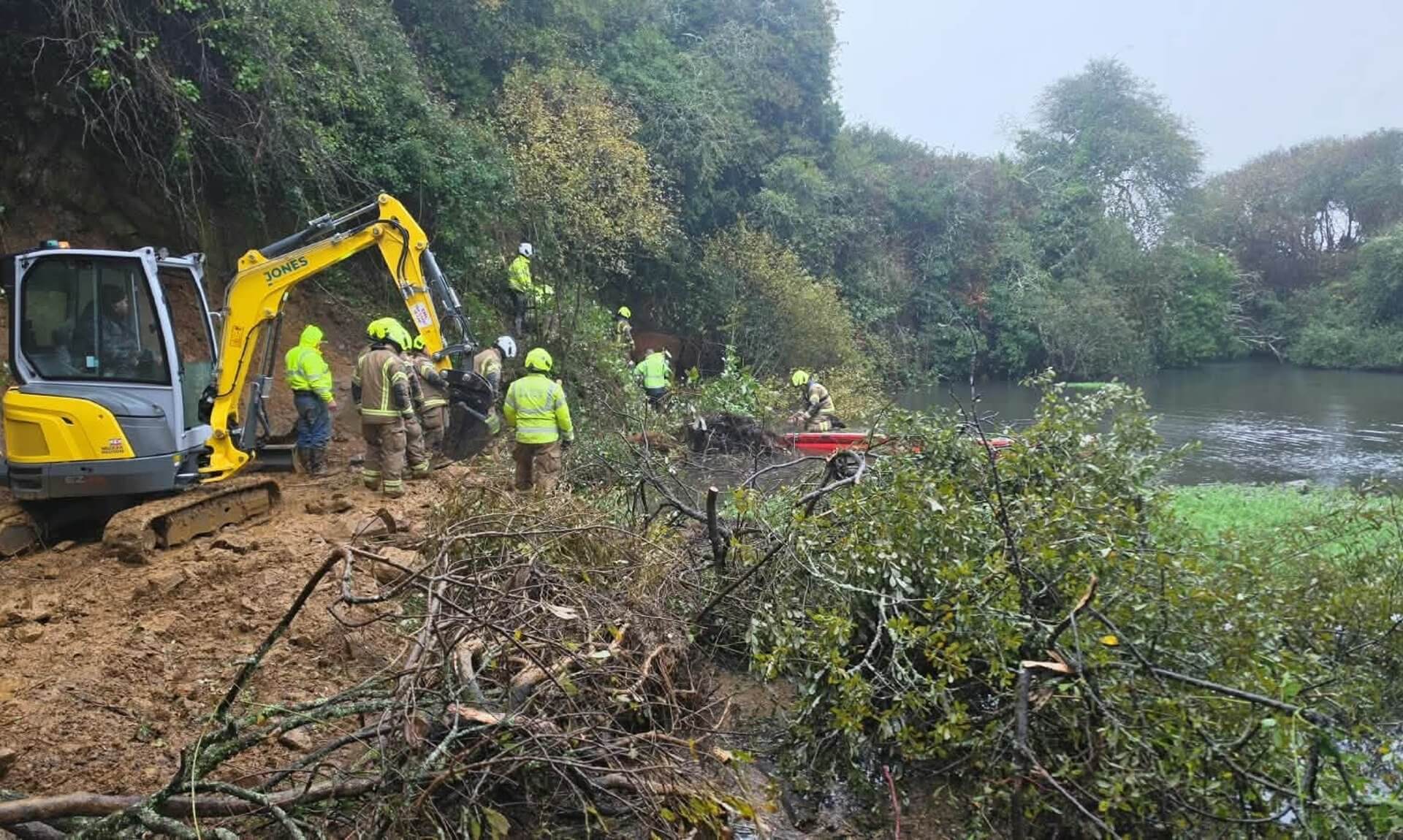 Firefighters and a mini digger clearing undergrowth beside water during a cow rescue