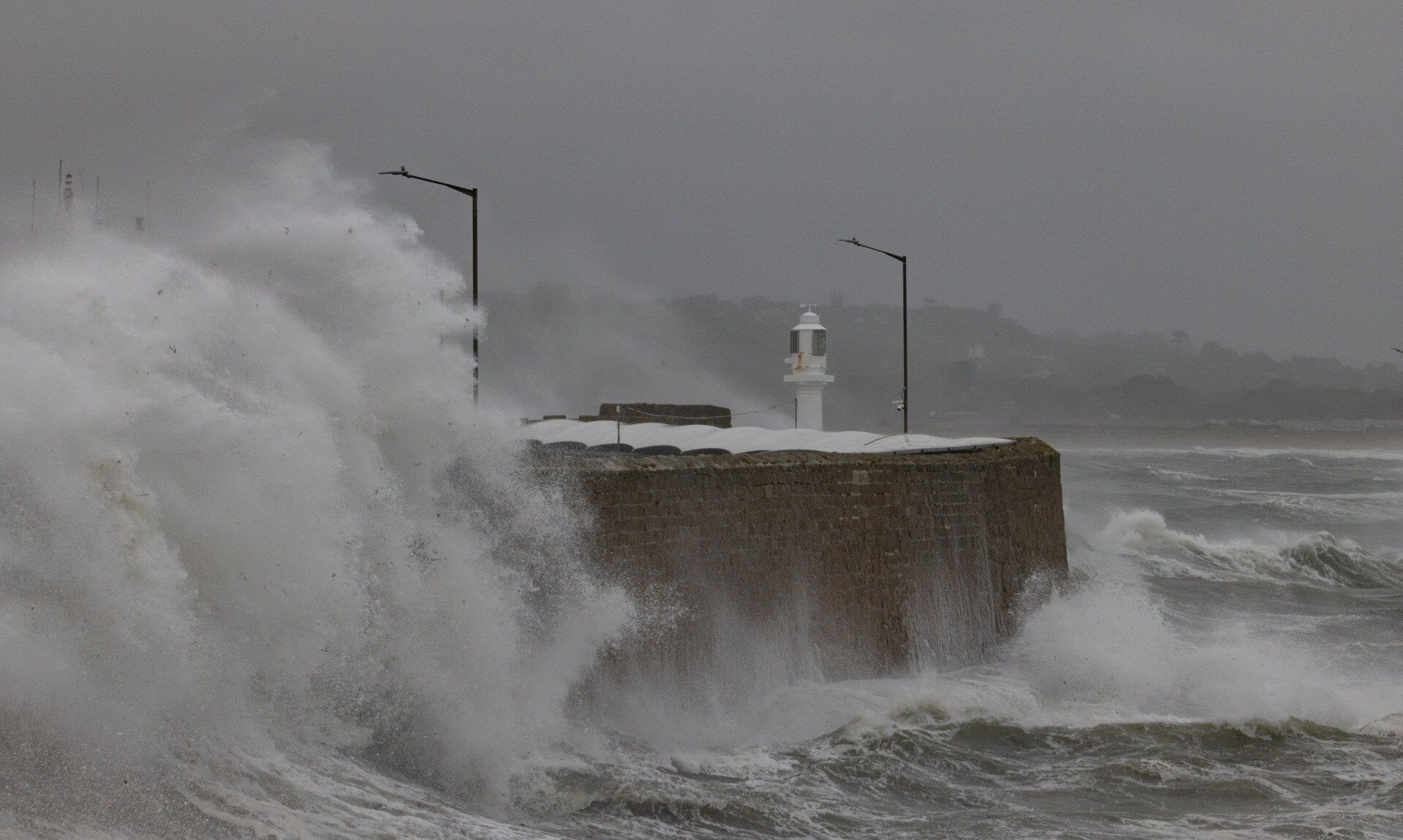 Storm waves crashing over a harbour wall and lighthouse in Cornwall