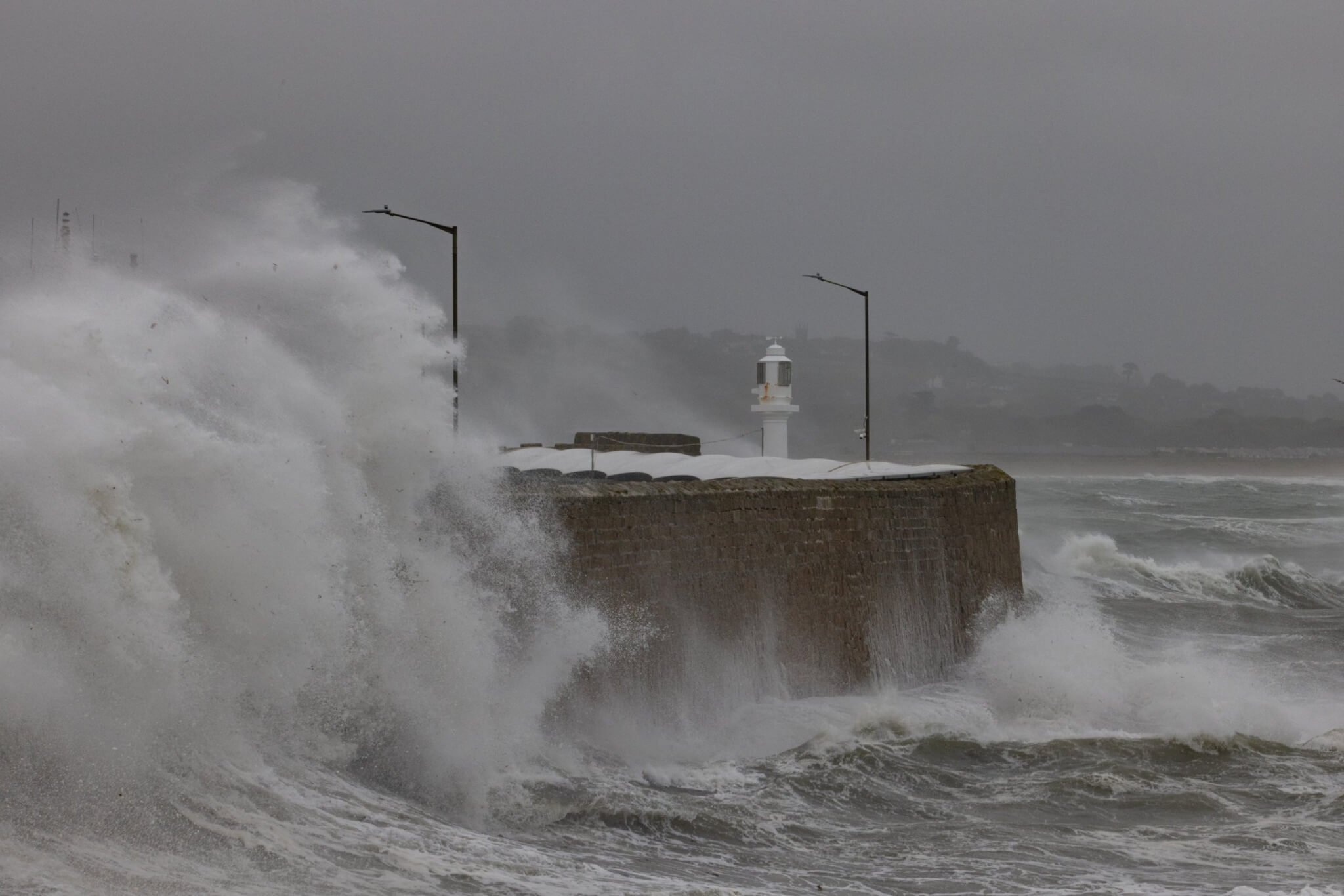 Storm waves crashing over a harbour wall and lighthouse in Cornwall