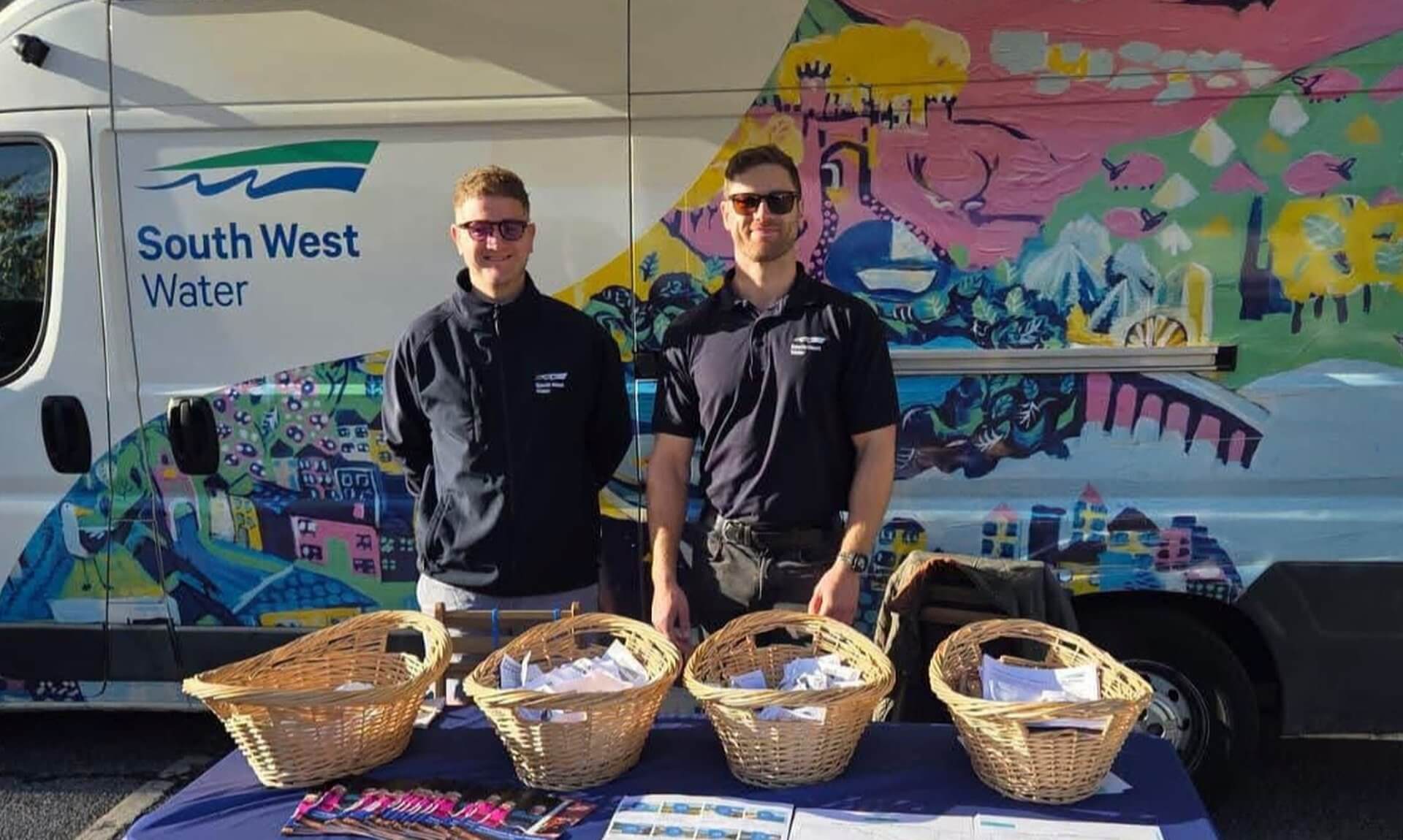Two South West Water staff at a community drop-in stand in front of a painted van